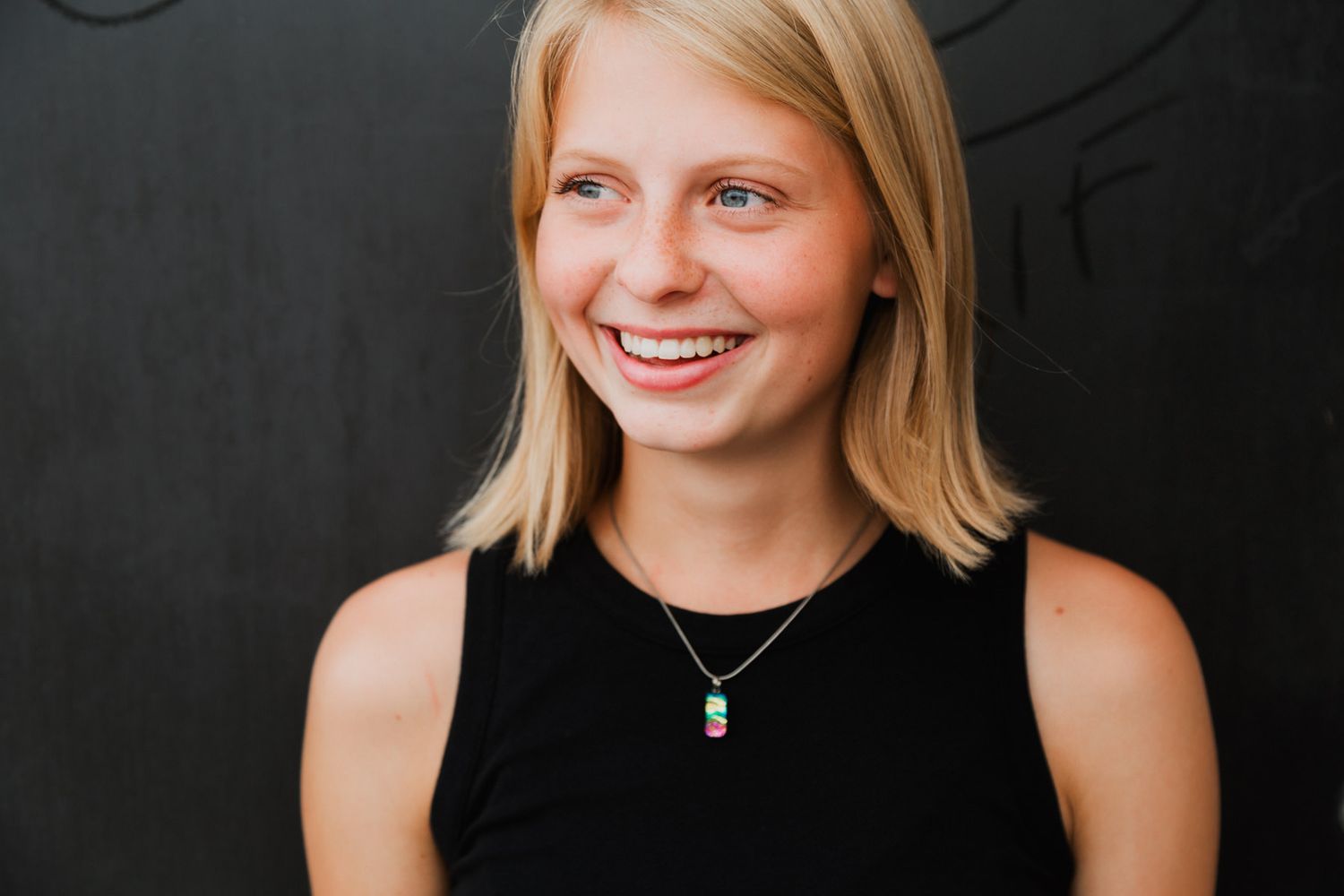 Smiling blonde teenage girl with blue eyes wearing a black sleeveless top and teal pendant necklace.