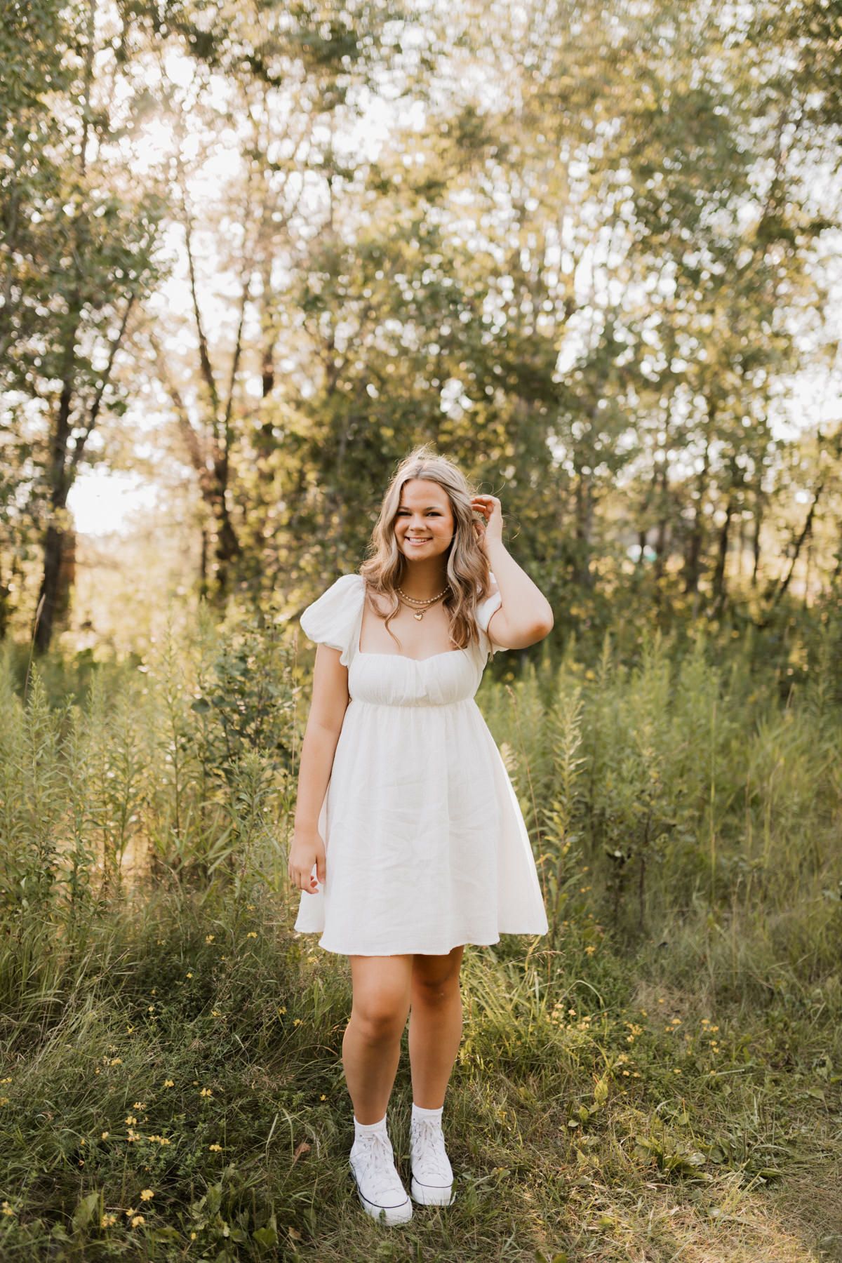 Young blonde woman in white dress and white sneakers smiling in a lush green forest setting.