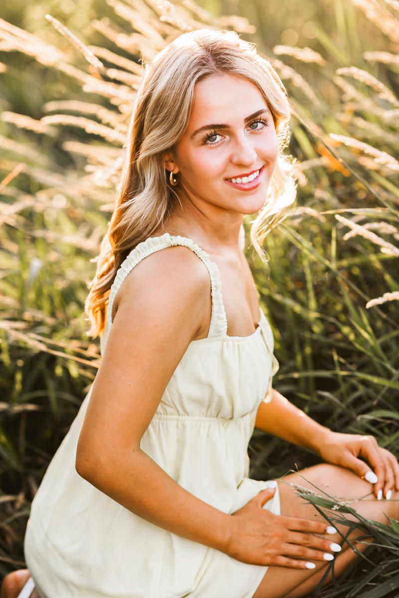 Young woman in white sundress sits gracefully in sunlit field of tall golden grass during a summer afternoon.