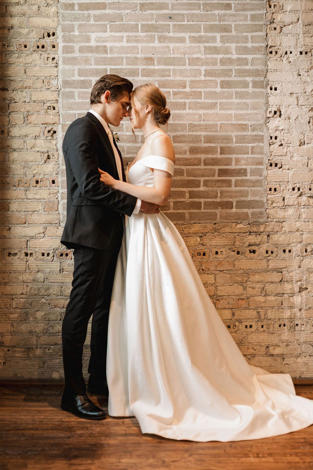 A couple shares an intimate moment in their wedding attire against a rustic brick wall backdrop.