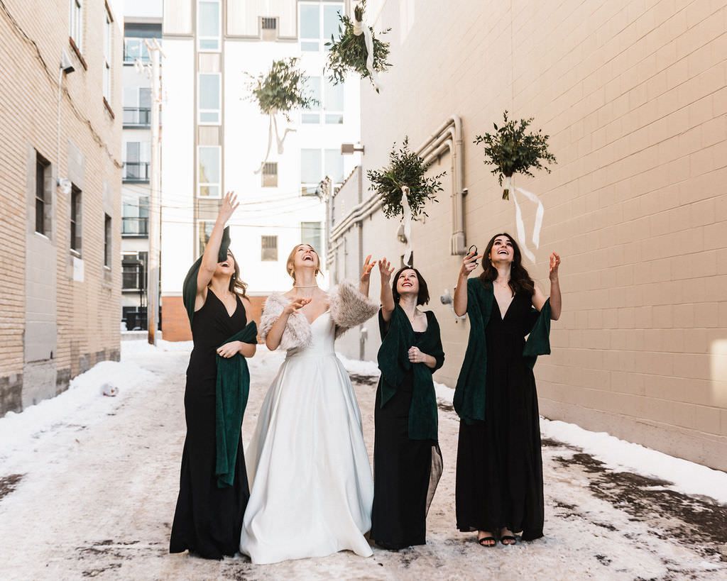 Bridal party in dark green dresses and white gown celebrate joyfully against light colored wall.