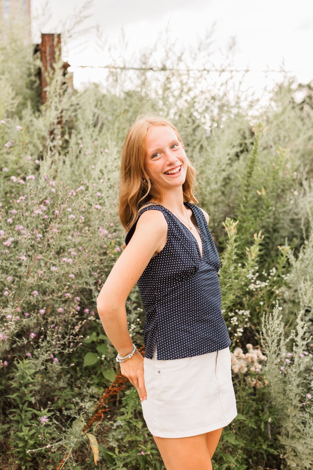 A person wearing a navy polka dot top poses joyfully in a garden setting with greenery and flowers.