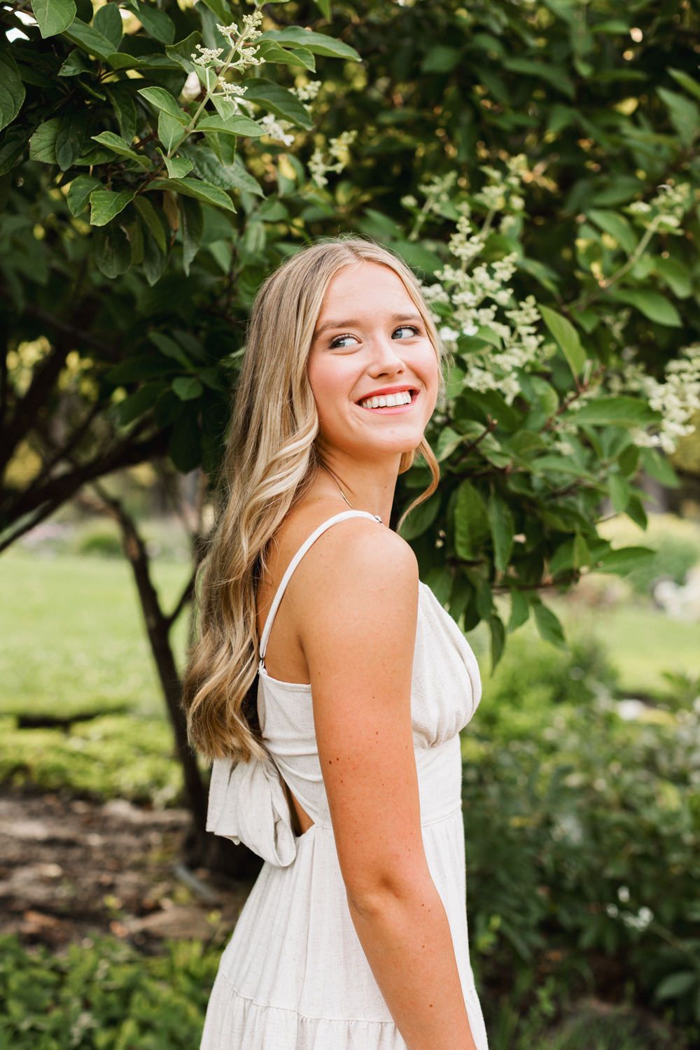 Portrait of someone in a white summer dress smiling while standing in a lush garden setting with flowering trees.