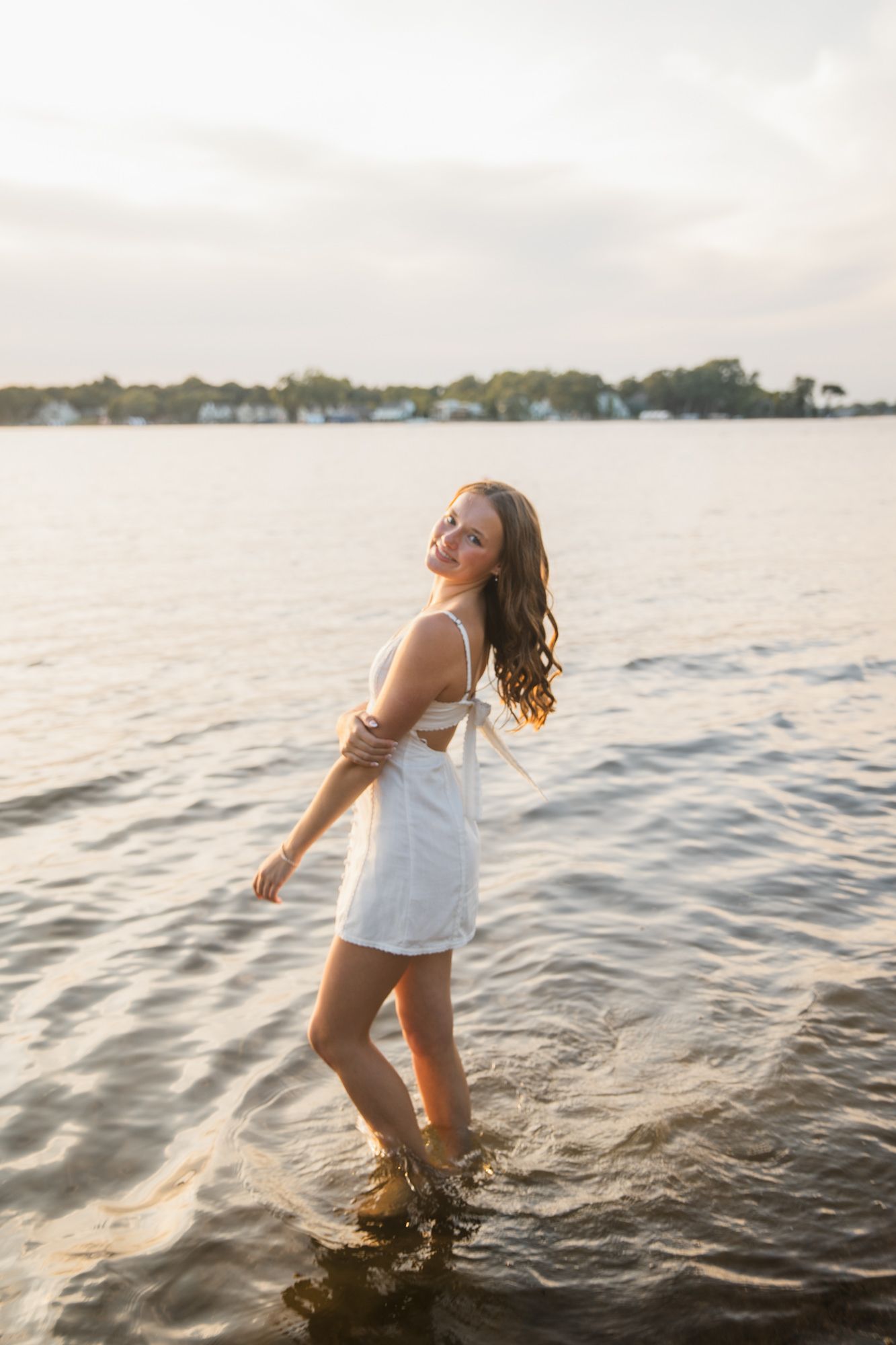 Young woman in white dress standing in shallow water at golden hour with scenic waterfront background.