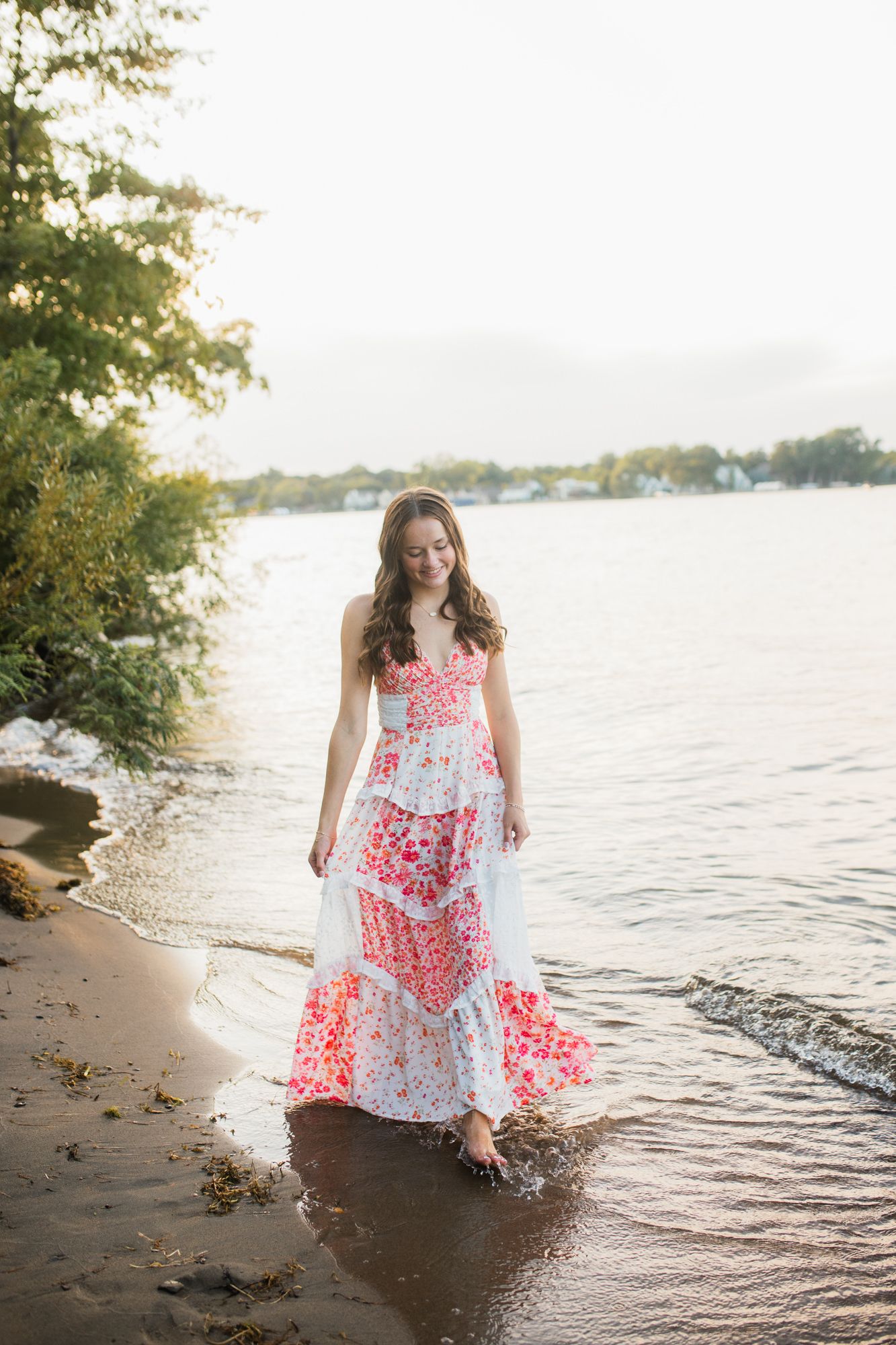 Woman in floral maxi dress wading in shallow lake water at sunset near a tree-lined shore.