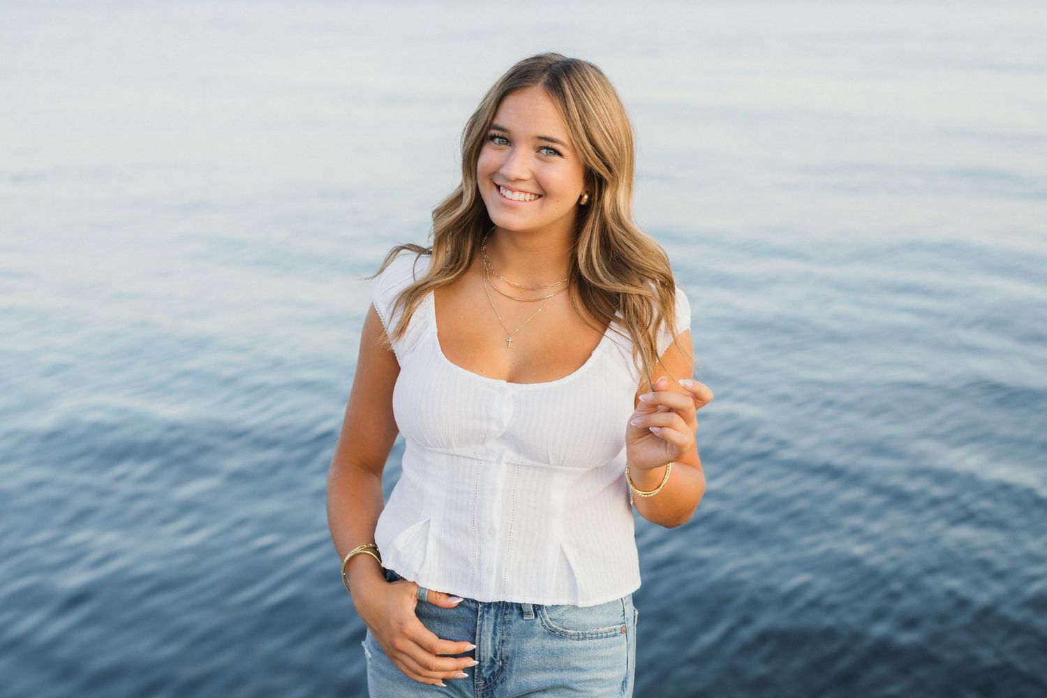 Young woman in white top and jeans smiling by the ocean on a sunny day.
