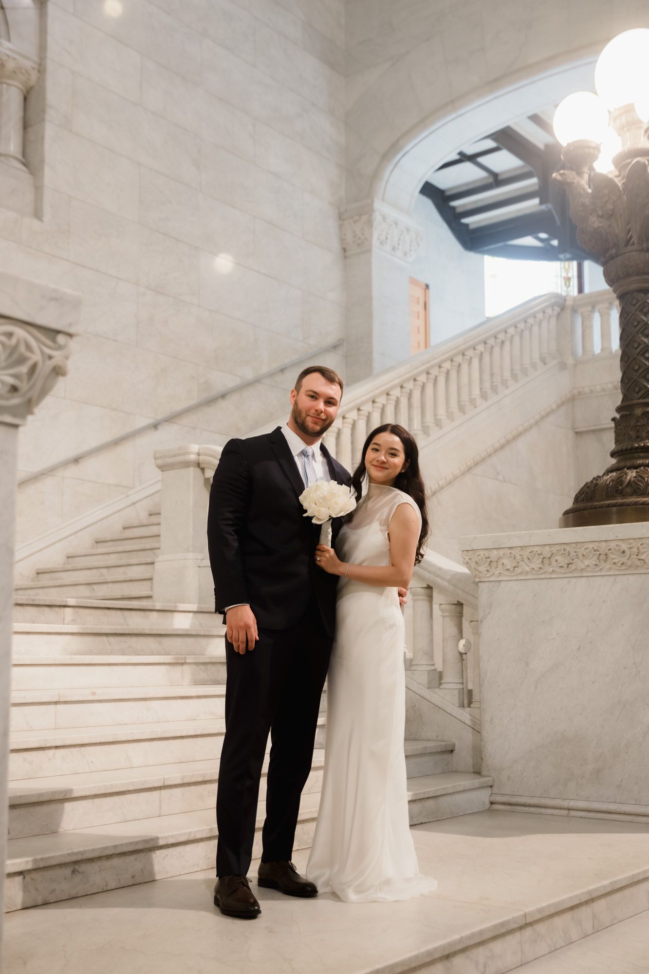 Bride and groom posing on marble staircase in elegant venue, bride holding white bouquet in wedding dress.