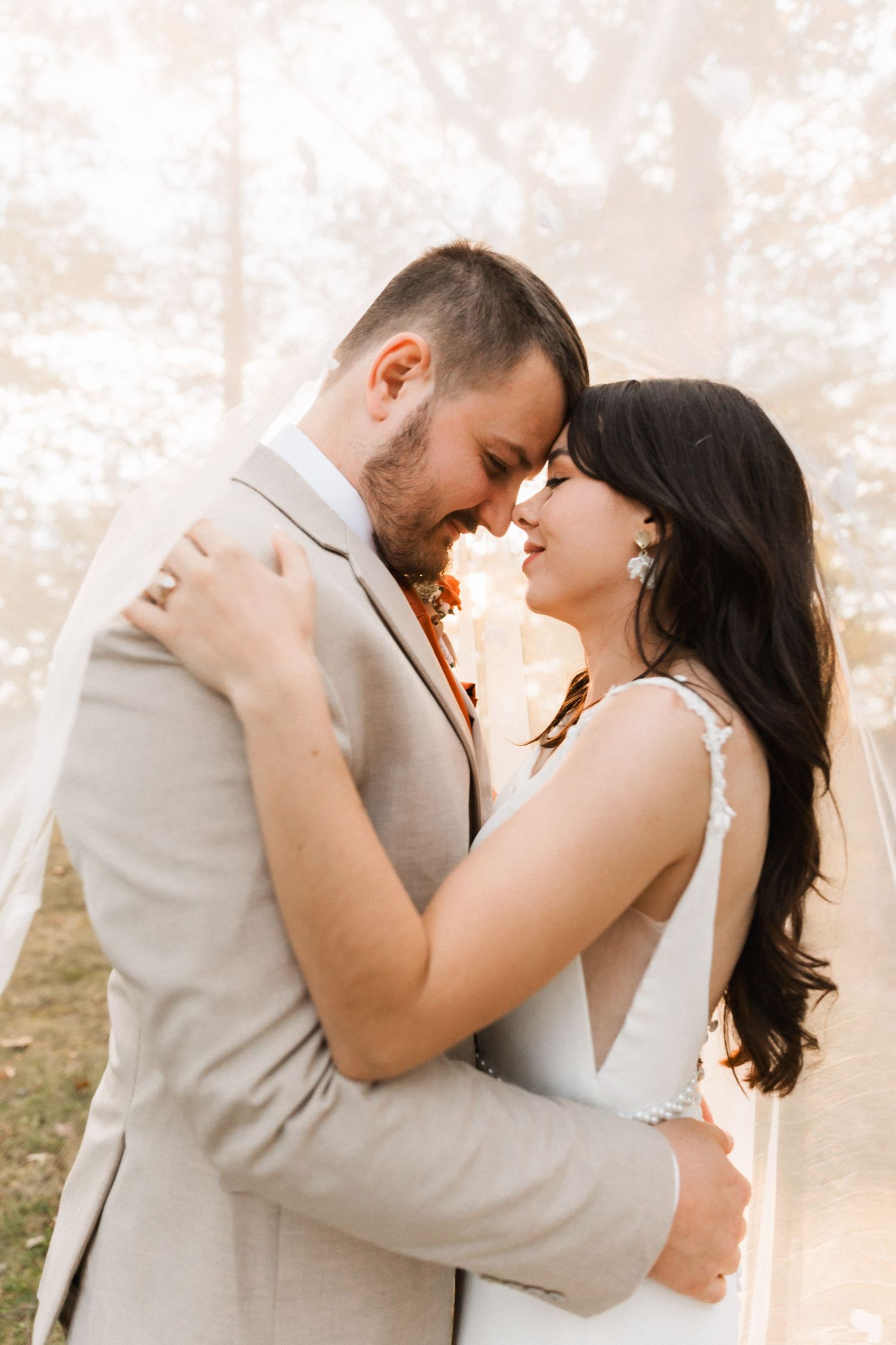 A romantic wedding couple embraces in soft sunlit outdoor portraits wearing grey suit and white dress.
