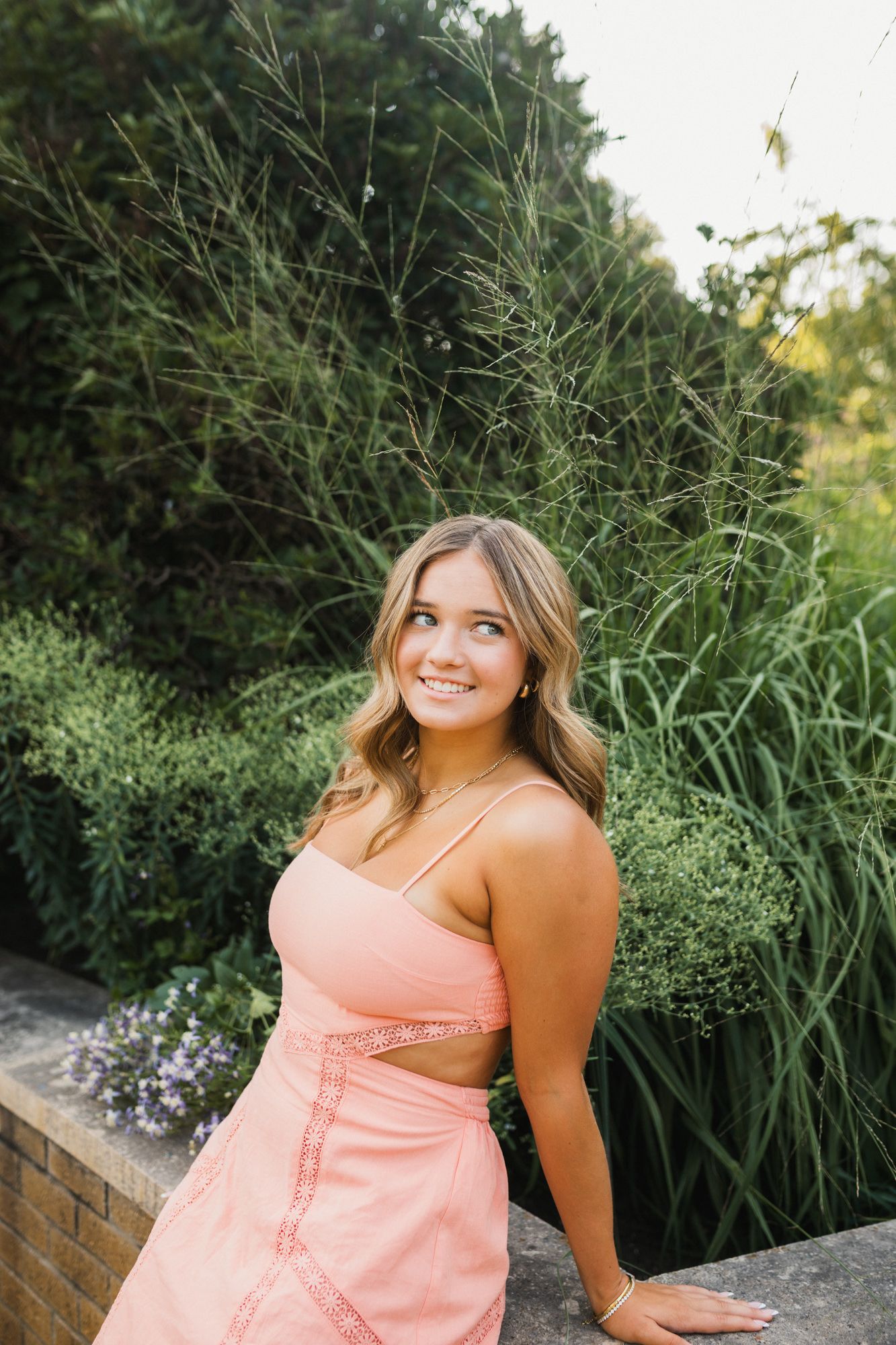 Young woman in pink dress smiling outdoors with lush green plants in background.