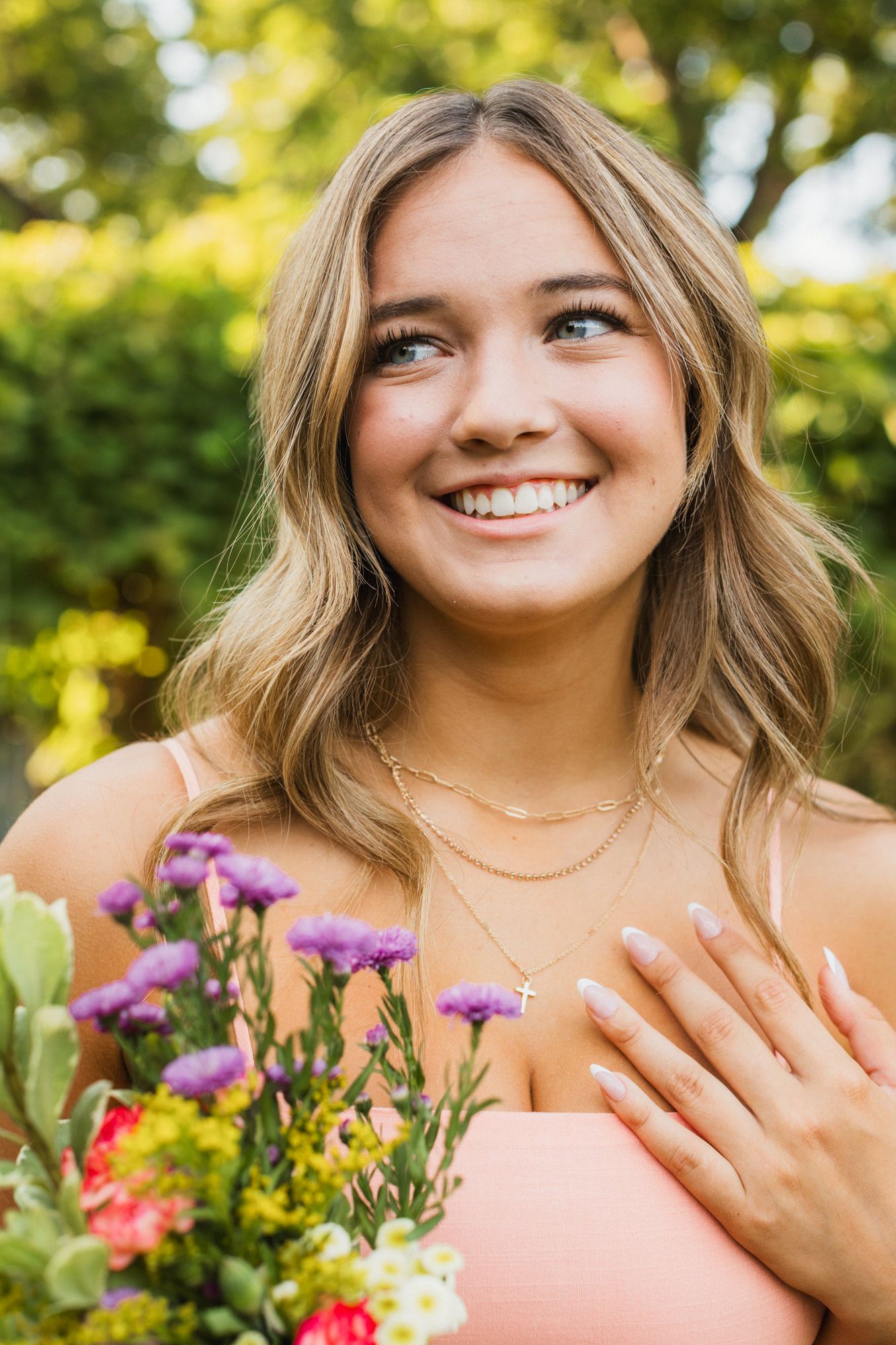 Smiling young woman with blonde hair holding colorful wildflowers outdoors, wearing a pink top and gold layered necklaces.