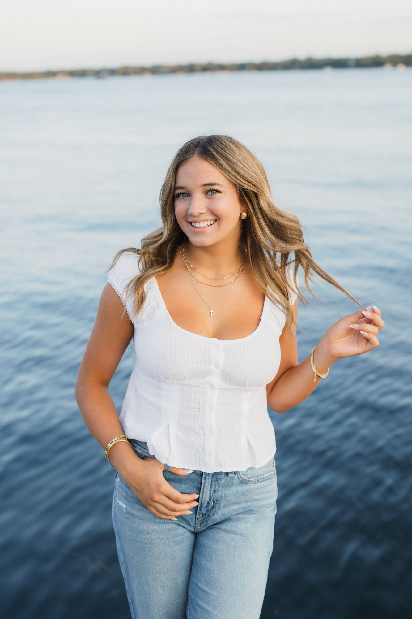 Smiling young woman in white top and jeans posing by a calm blue lake on a sunny day.