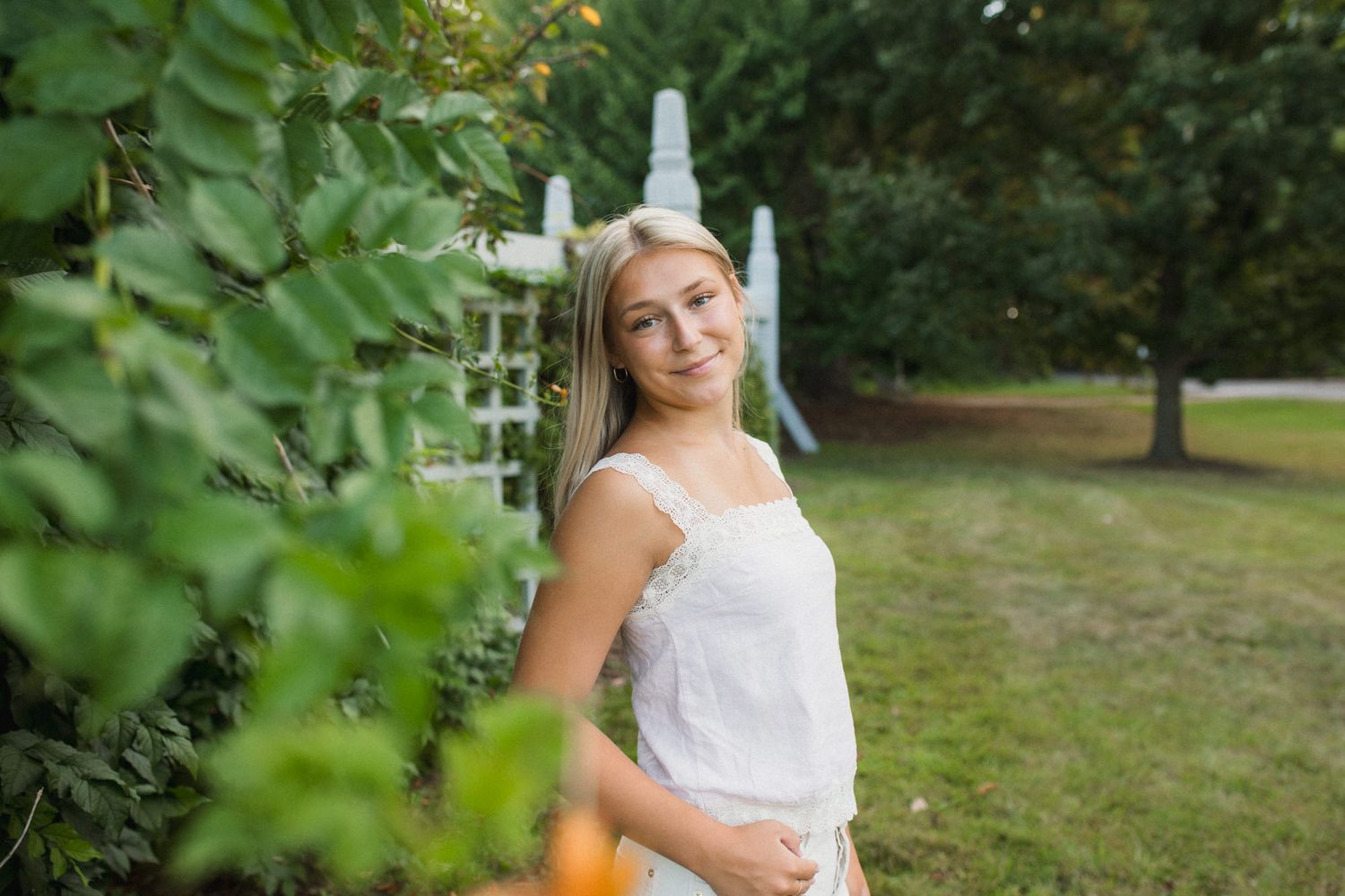 Young blonde woman in white dress smiling outdoors among lush green trees and garden arbor.
