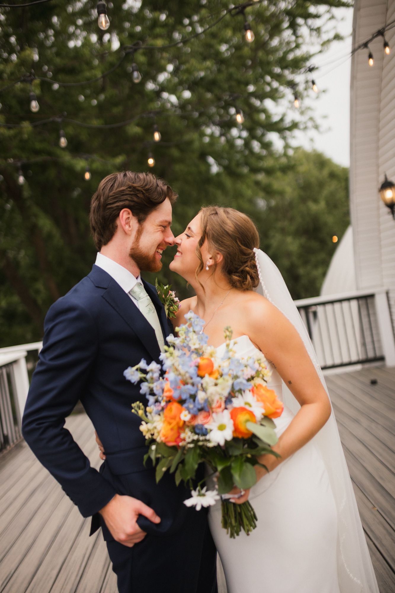 Bride and groom share a romantic moment on a deck, bride holding a colorful bouquet of orange, blue, and white flowers.