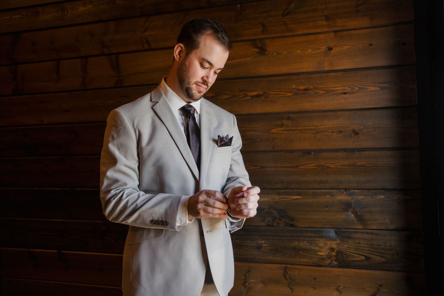 Groom in a light gray suit adjusts his cuff while standing against a rustic wooden plank wall.