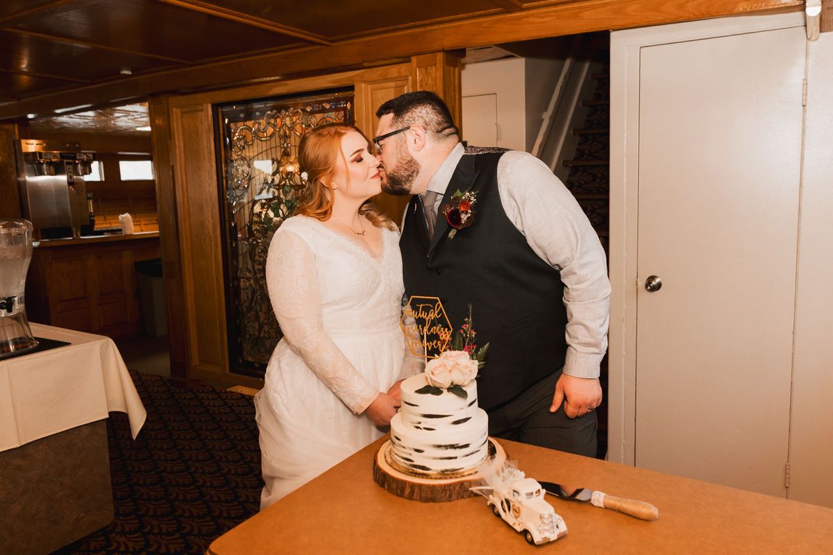 Wedding cake cutting celebration in warm-lit wooden interior of boat.