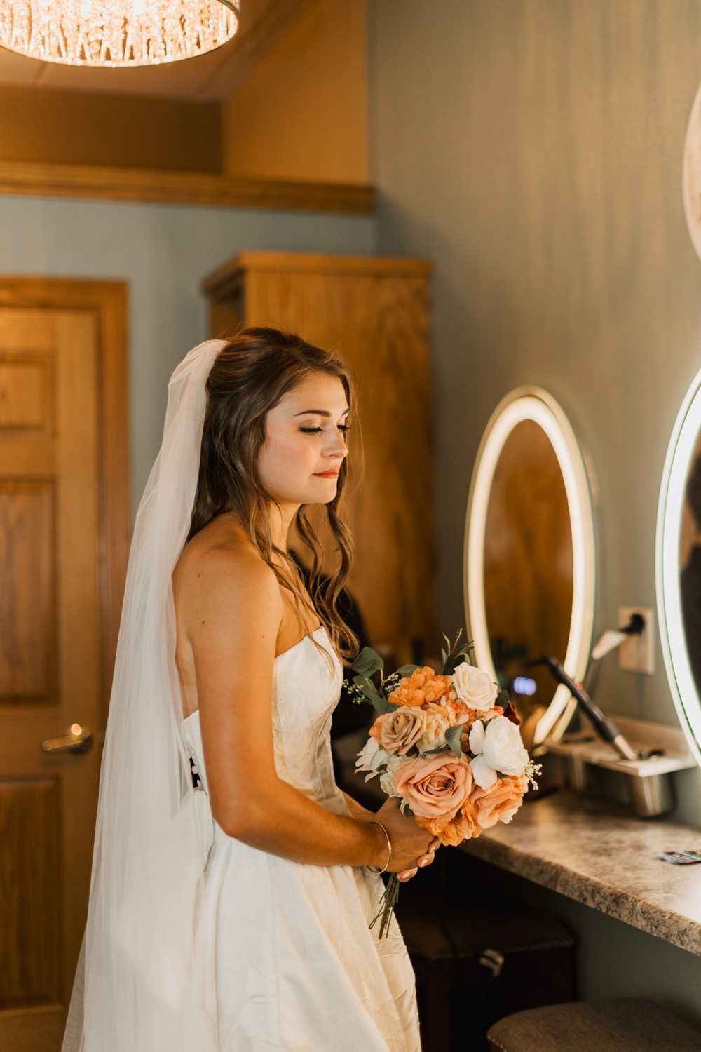 Bride holding a peach and white bouquet while getting ready in a warm-lit room.
