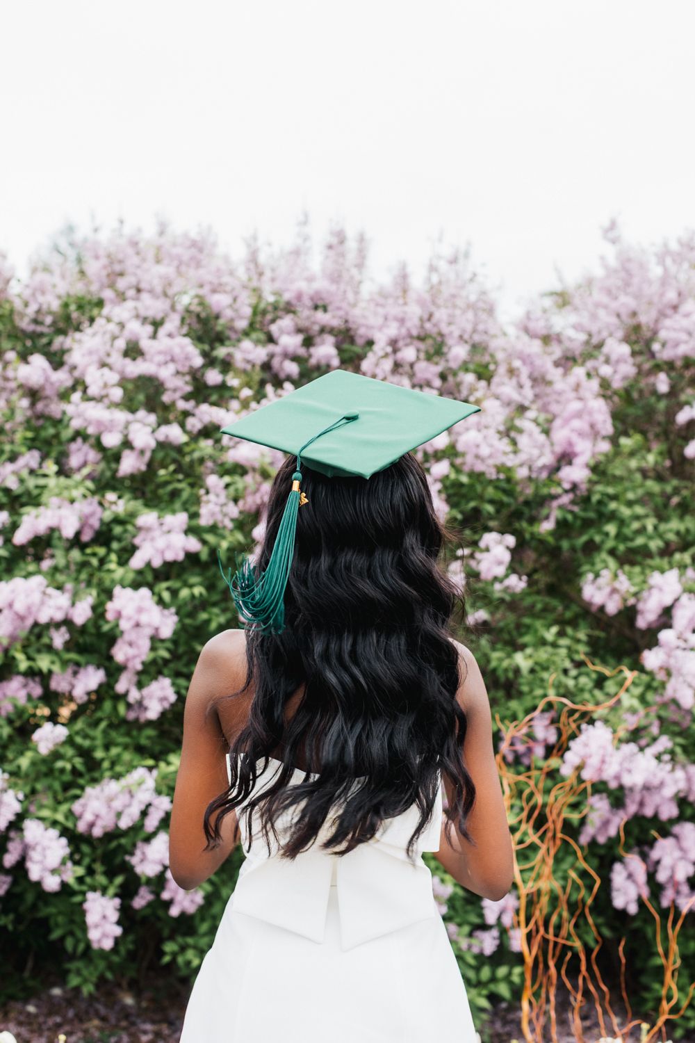 Back view of graduate with long wavy hair wearing green cap against blooming pink flowers.