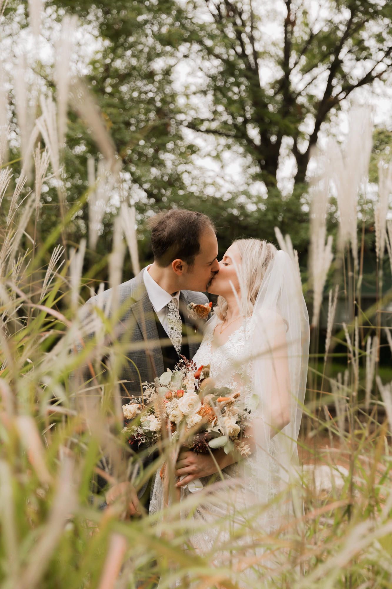 A bride and groom share a romantic kiss surrounded by tall ornamental grasses, bride holding a boho floral bouquet.