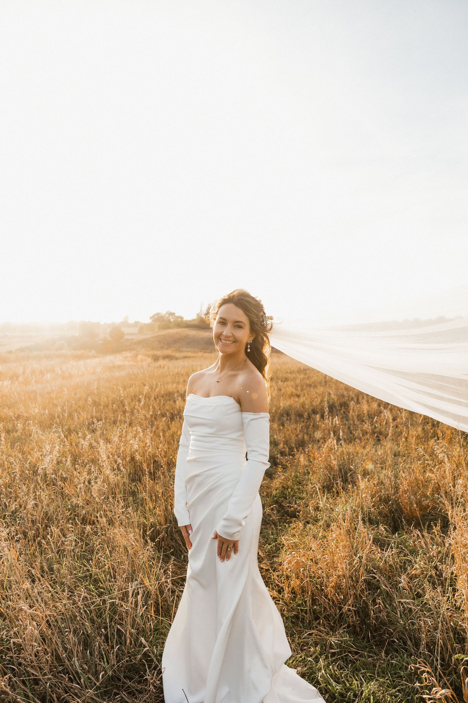 Smiling bride in an off-shoulder white wedding gown stands in a golden grass field at sunset as her veil blows in the wind.
