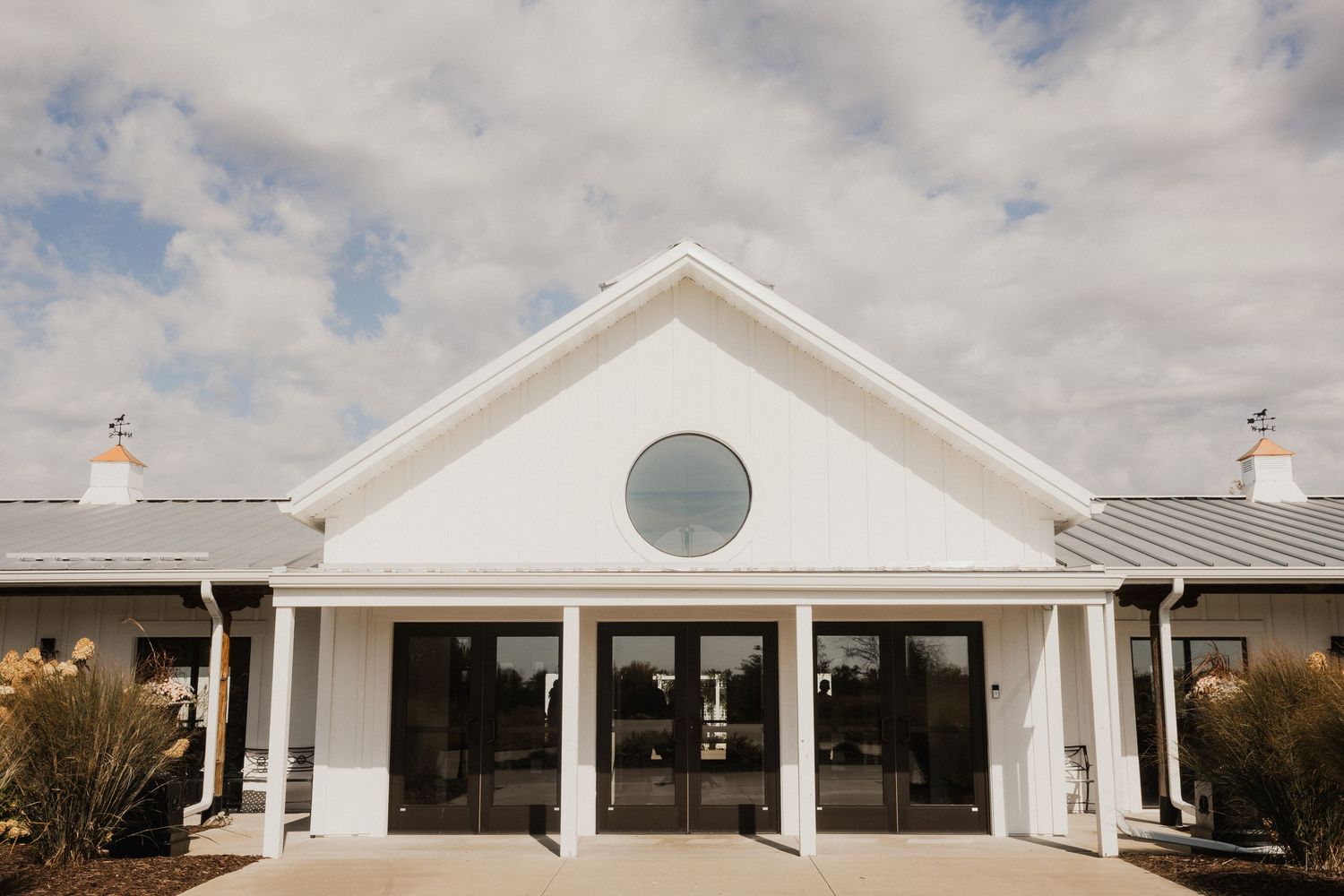 White event venue building with a gabled roof, round window, and glass double doors under a partly cloudy blue sky.