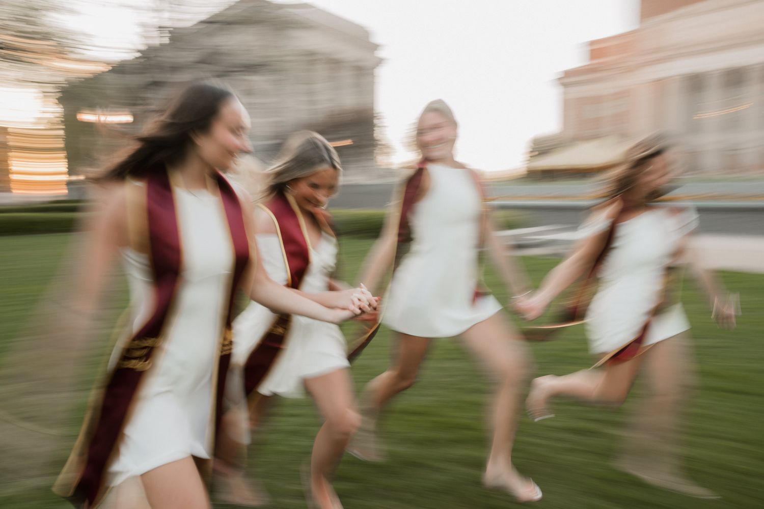 Motion-blurred photo of four women in white dresses and maroon graduation stoles running and holding hands.