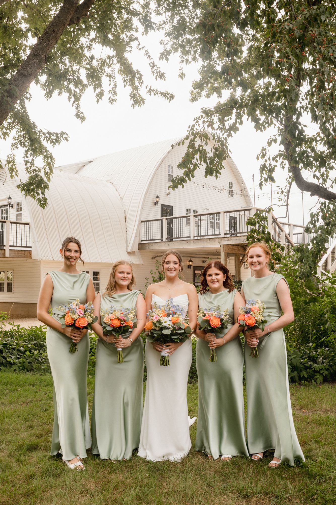 Bride in white gown with four bridesmaids in sage green dresses holding colorful bouquets outdoors at a wedding venue.