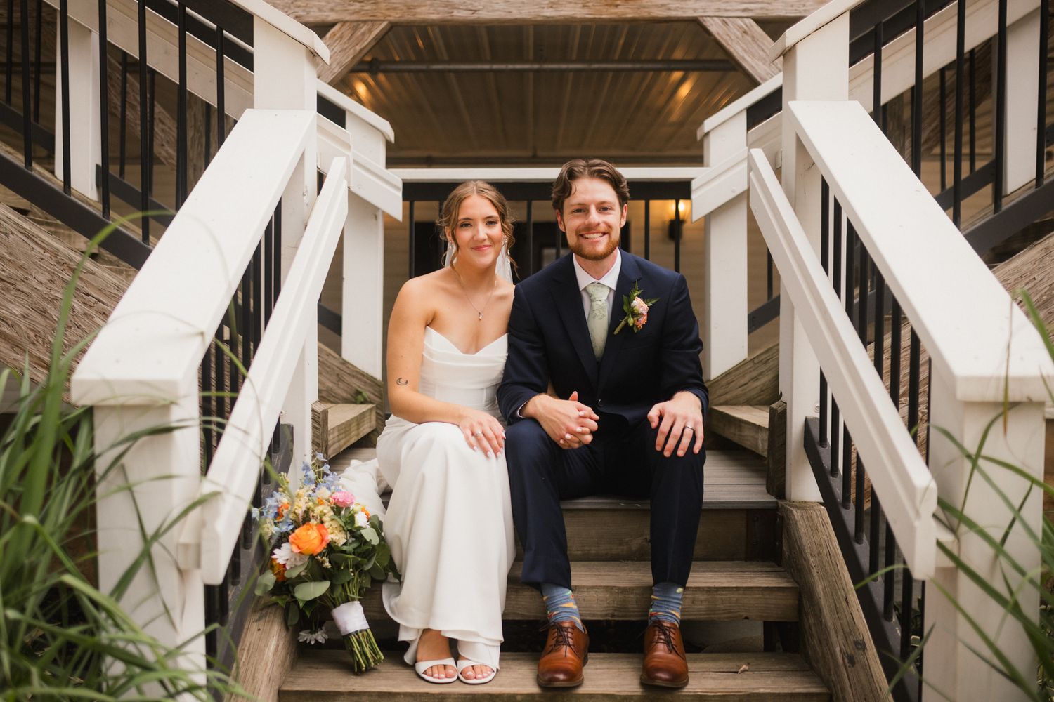 Bride and groom sitting together on wooden steps, smiling, with a colorful bouquet, bride in white gown, groom in navy suit.