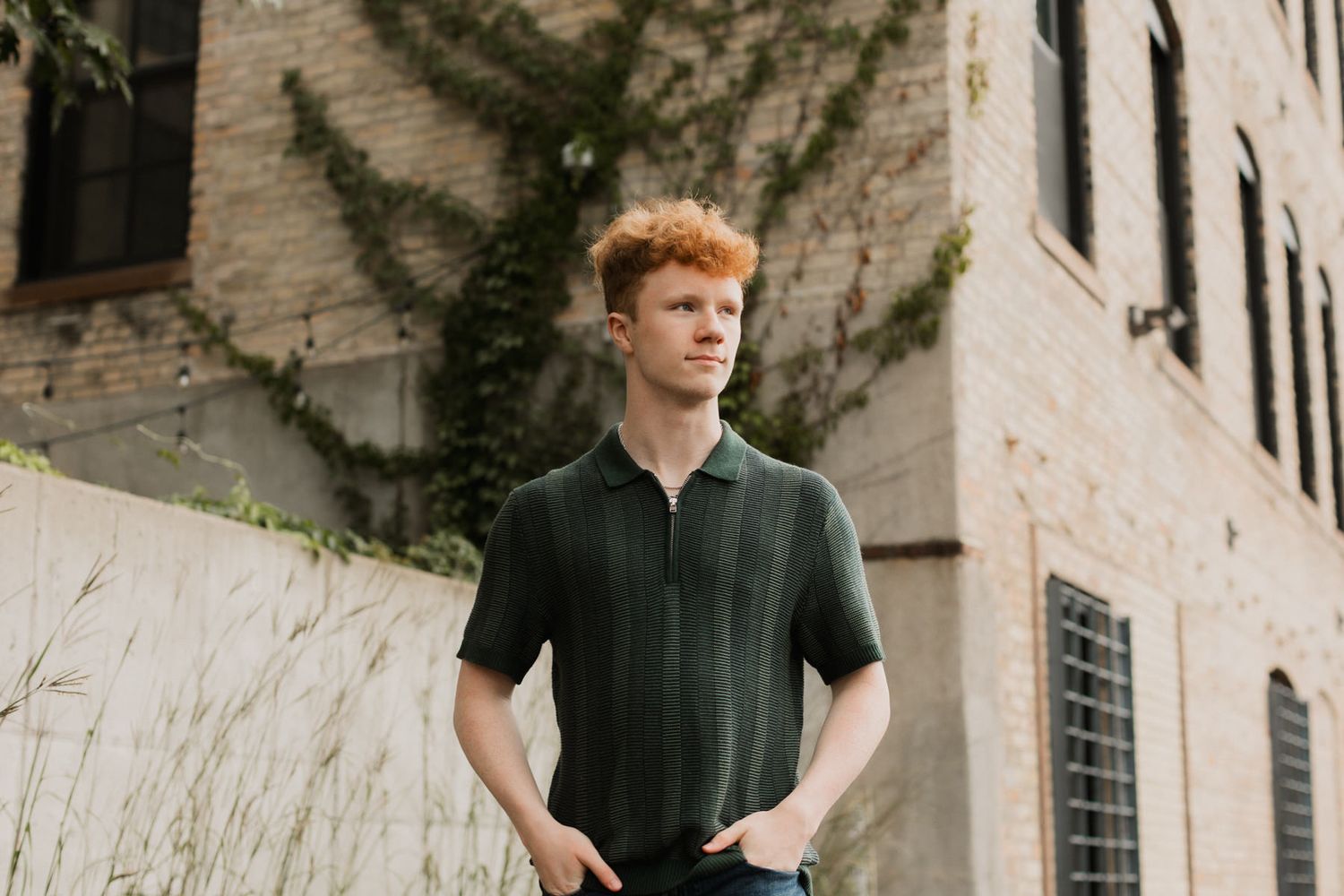 Young man with curly red hair wearing dark green knit polo shirt standing near ivy-covered brick wall.