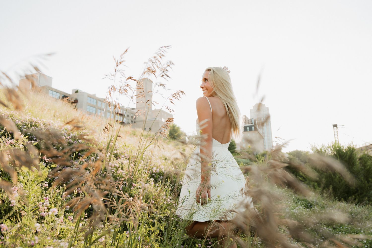 Blonde woman in white backless dress standing in golden field with tall grass and buildings behind her.