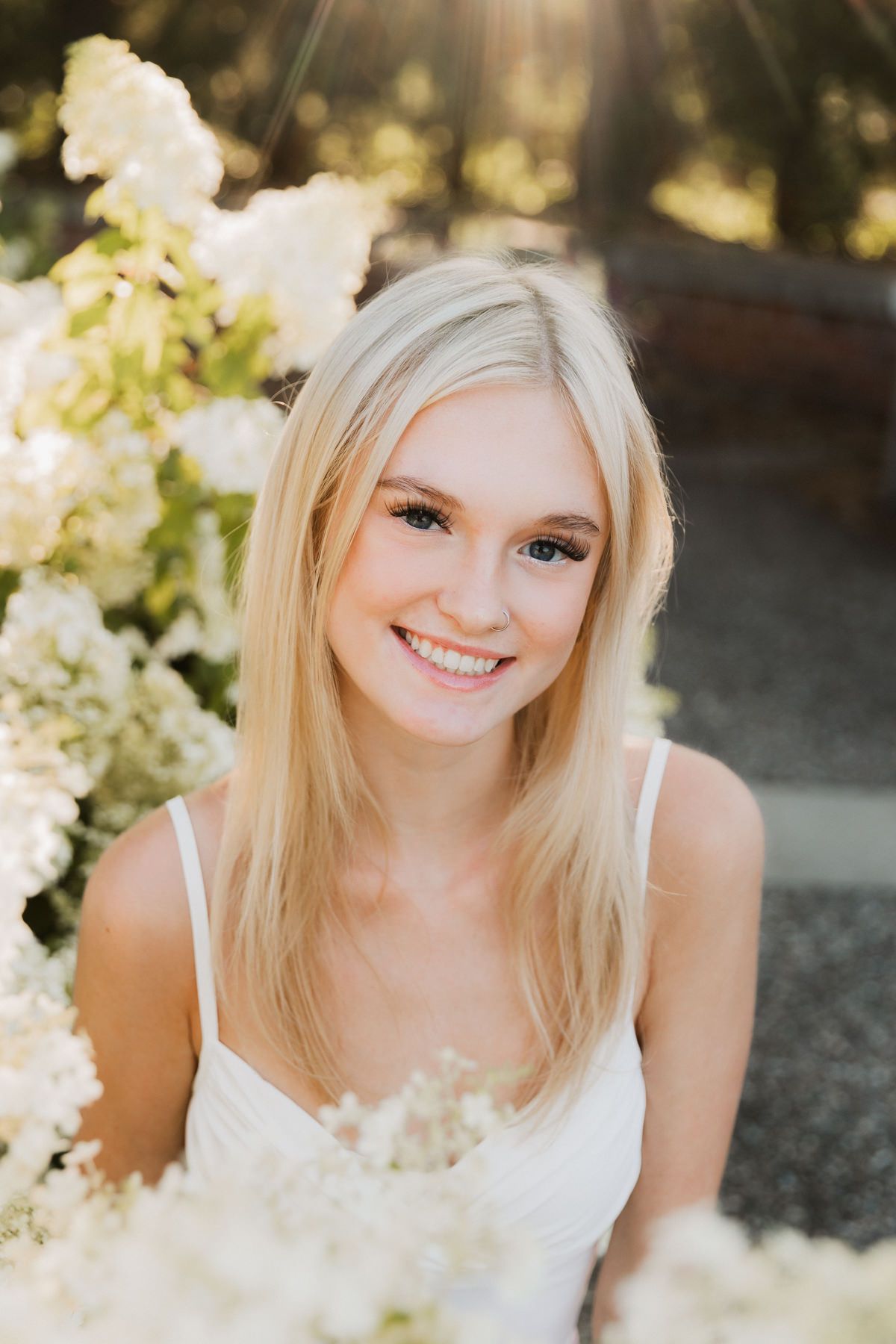 Smiling blonde teen girl in white dress surrounded by white hydrangea flowers in soft sunlight.