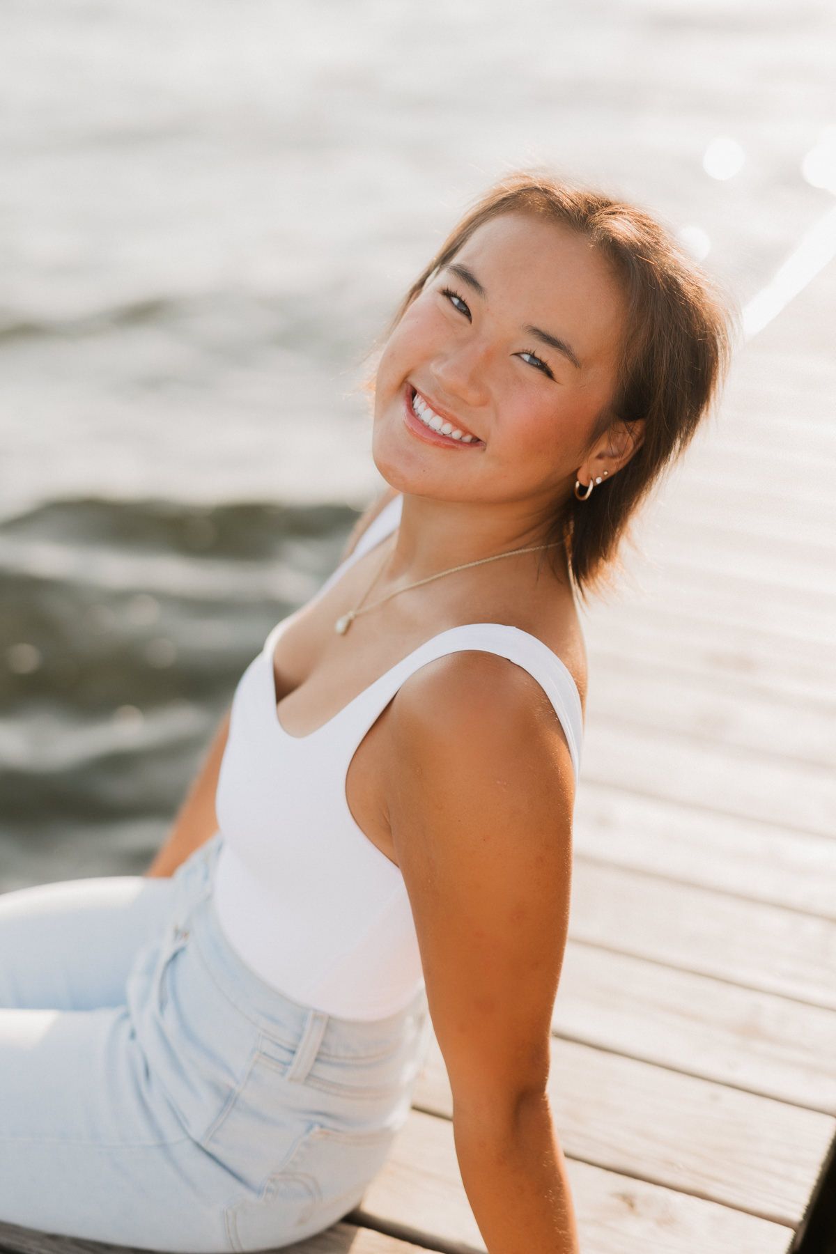 Young woman smiling on a wooden dock by the water, wearing a white top and light jeans in warm sunlight.