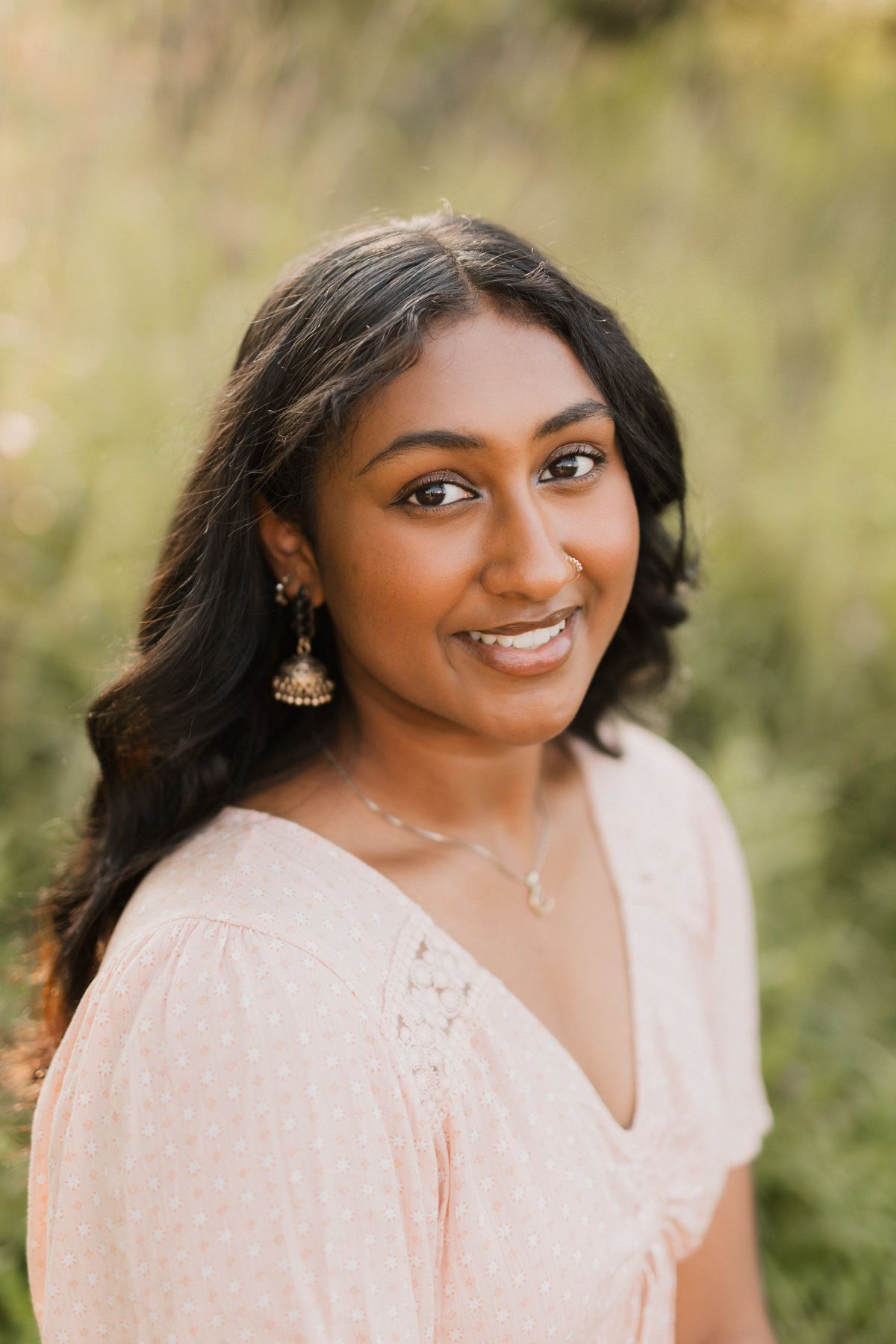 Young South Asian woman smiling outdoors, wearing a pink dress and gold jhumka earrings, with lush greenery in background.