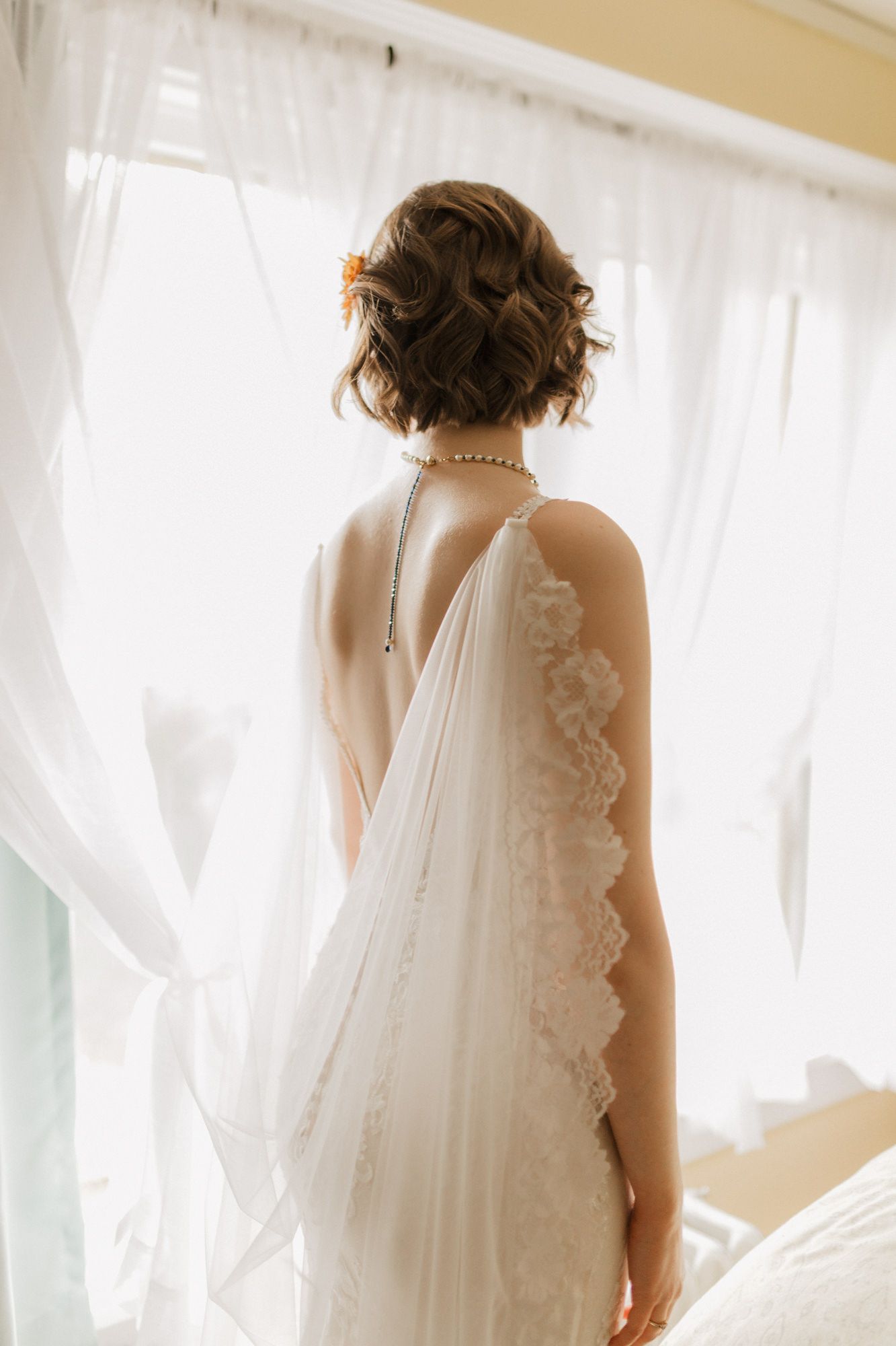 Bride in elegant white lace wedding gown with open back, pearl necklace, and updo hairstyle standing before sheer curtains.