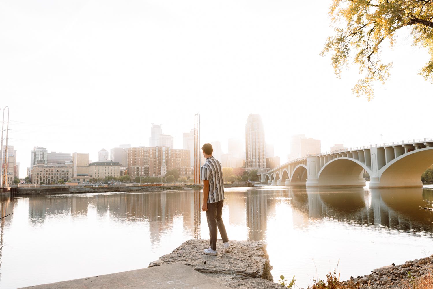 Person standing on riverbank during misty golden hour with city skyline and bridge in background.