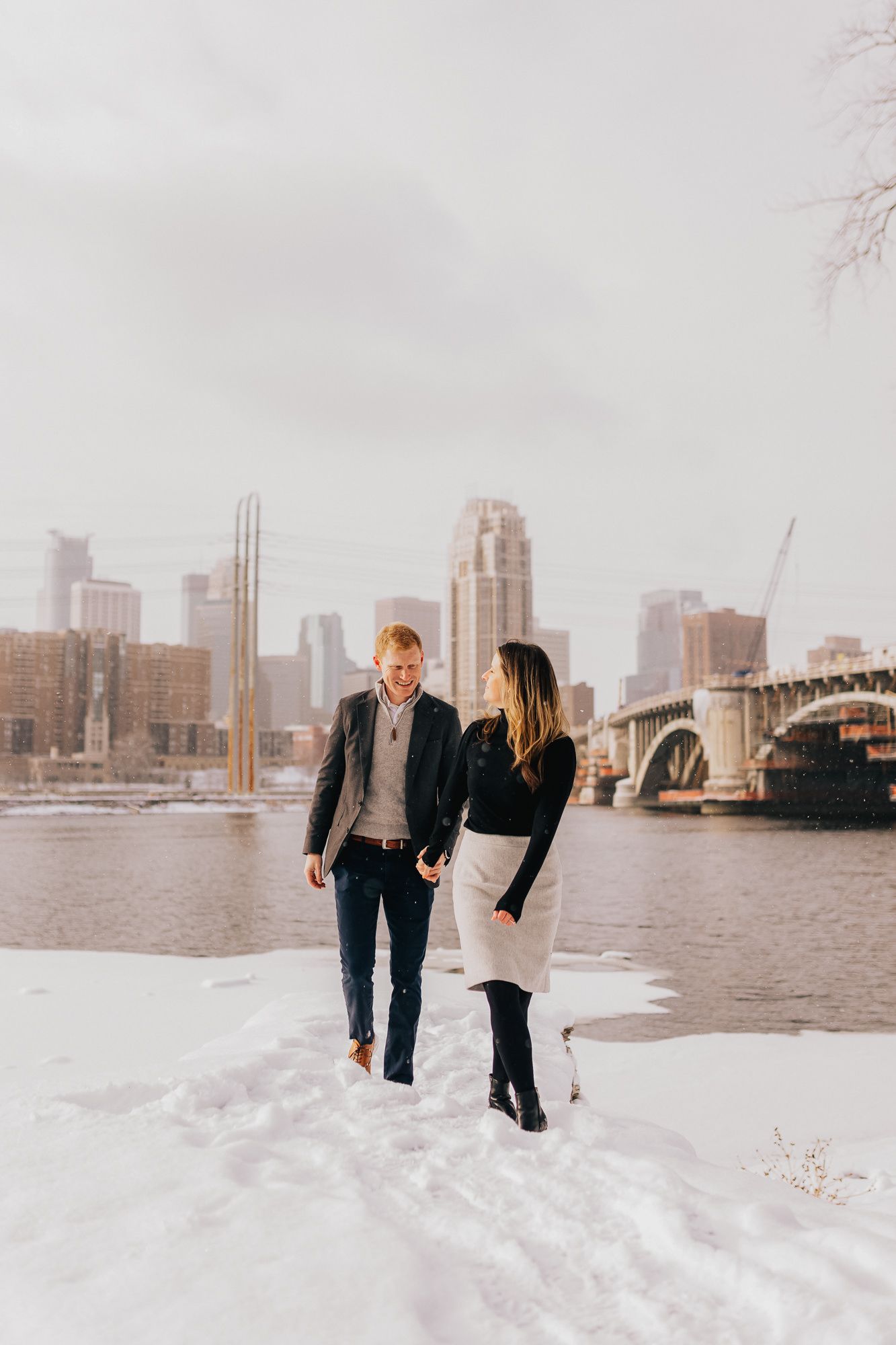 Couple walking hand in hand in snow by a river with a city skyline and bridge in the background.