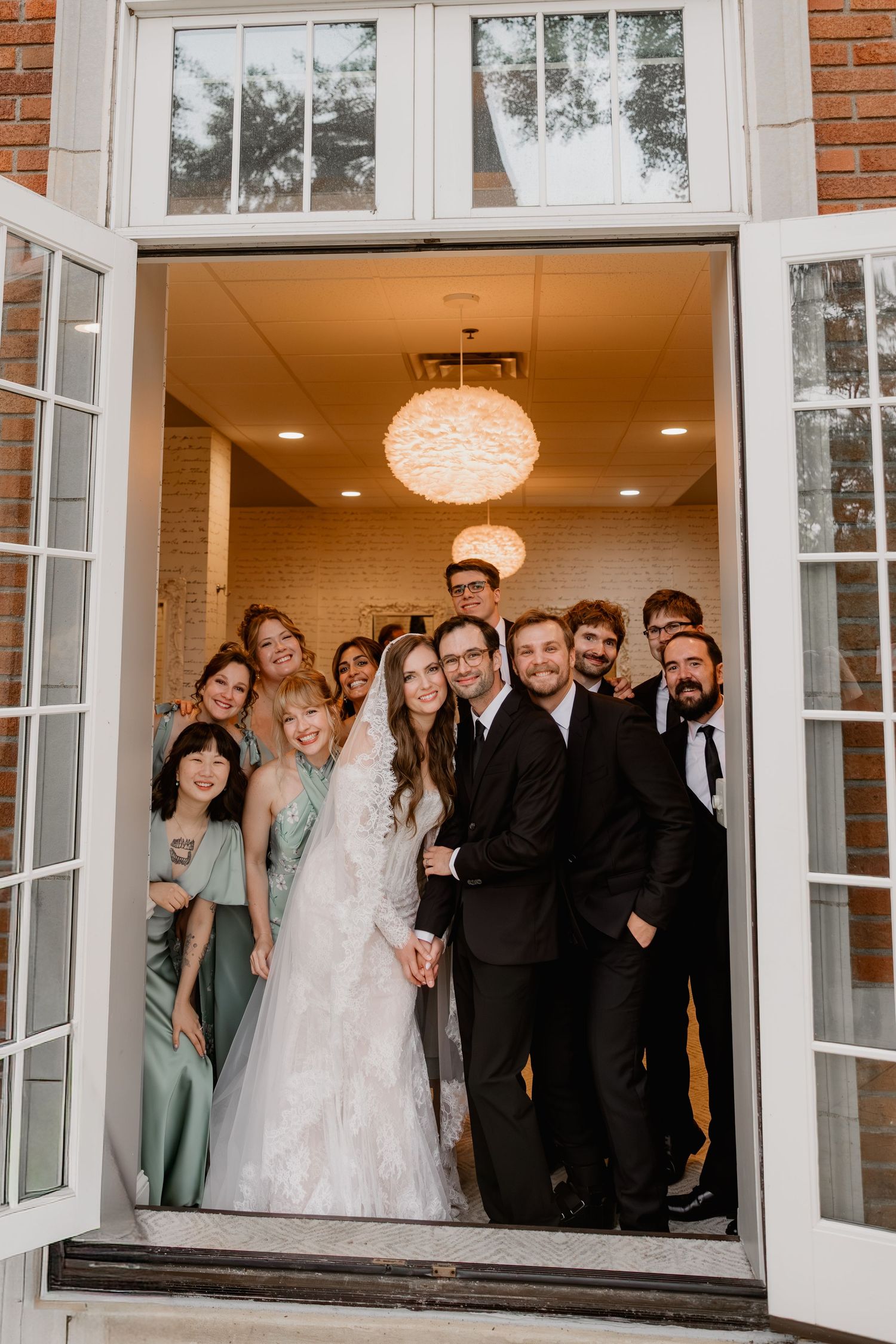 Wedding party poses in doorway, bride in white gown with bridesmaids in sage green and groomsmen in black suits.