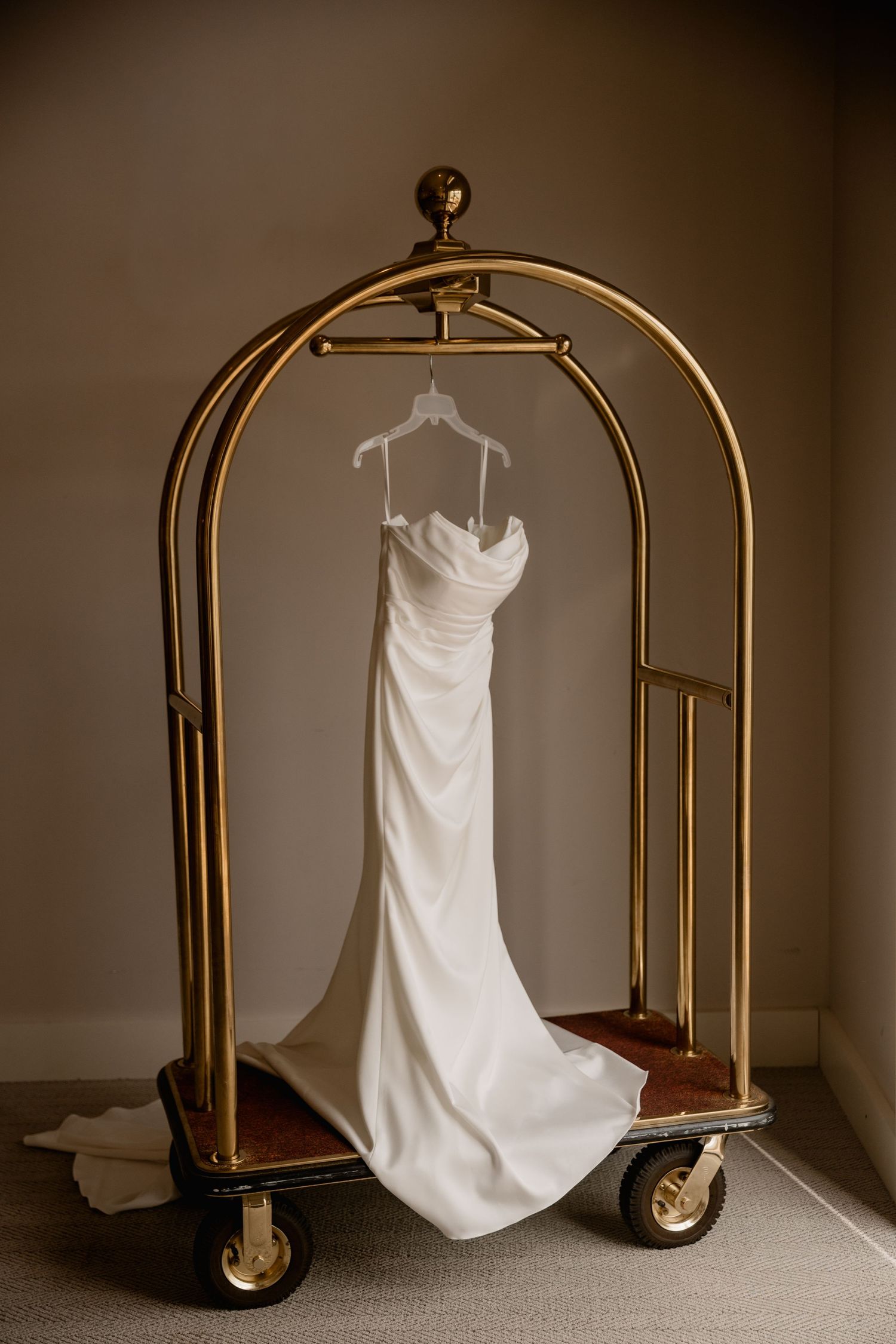 Elegant white wedding dress hanging on a gold hotel luggage cart in a neutral-toned room.