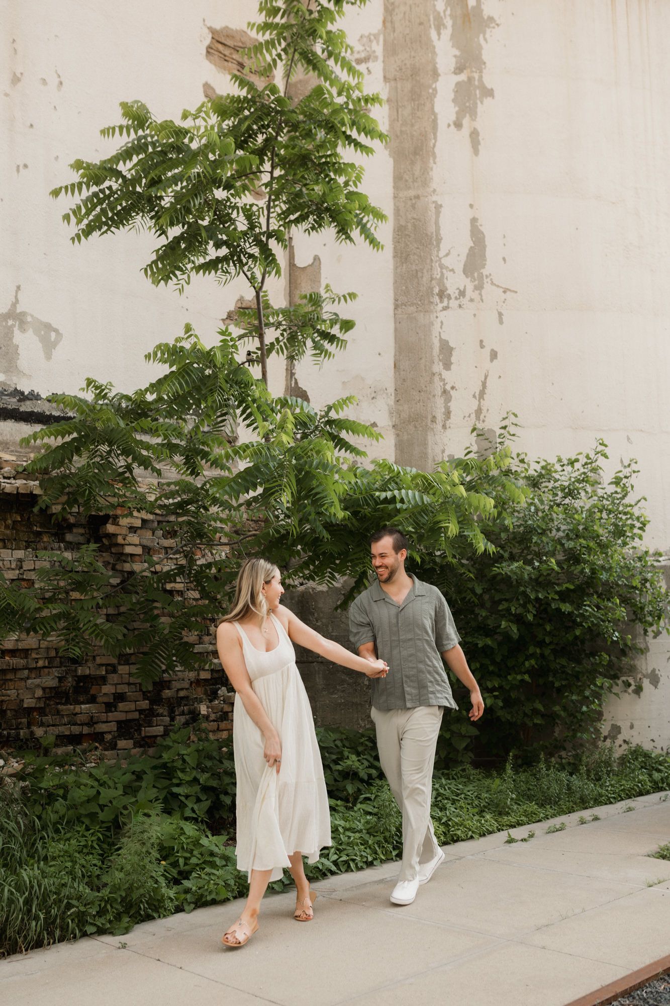 Couple walking hand in hand on a path surrounded by lush greenery and a rustic wall backdrop.