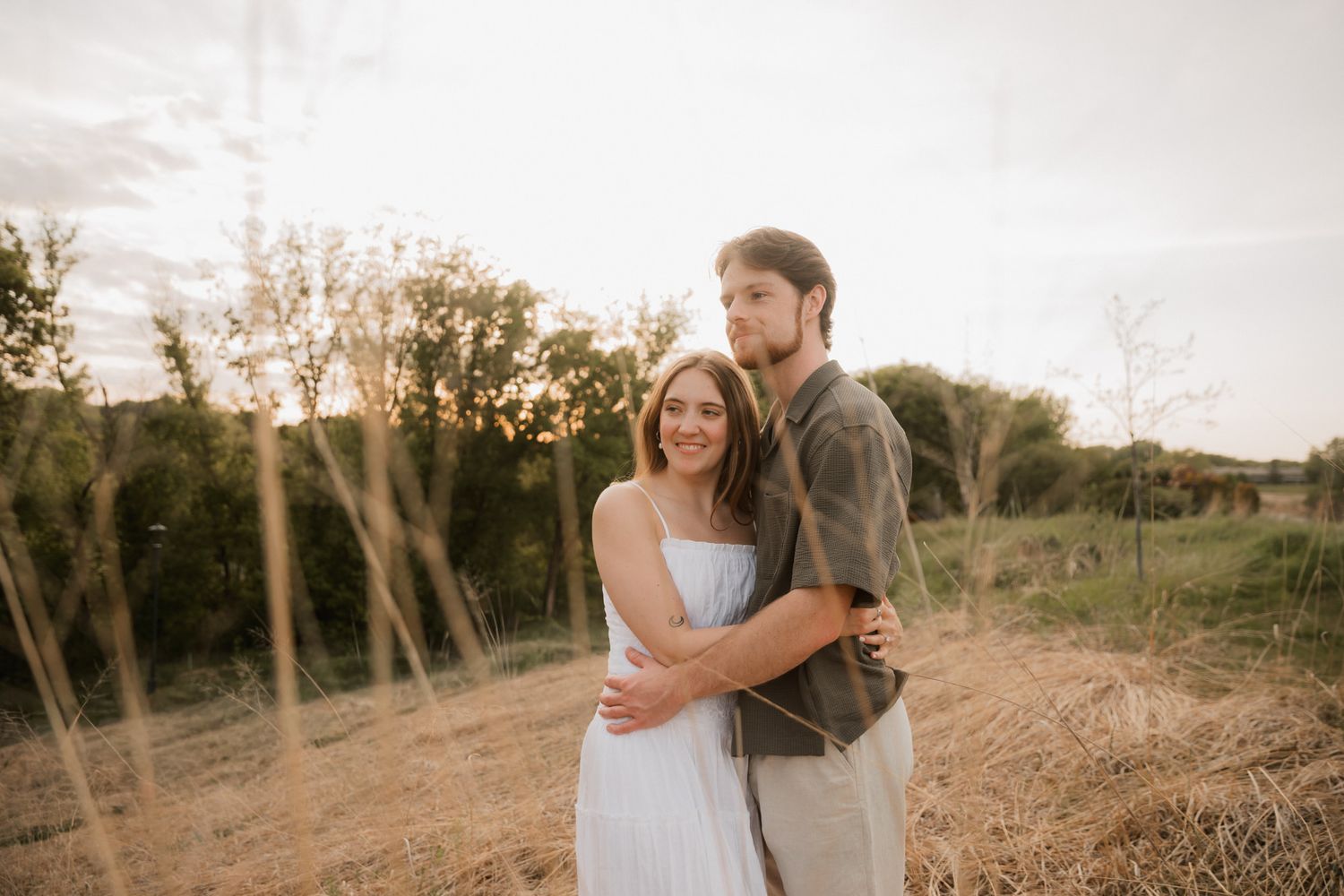 Couple embracing at golden hour in a field, woman in white dress smiling, man in olive jacket, lush greenery background.