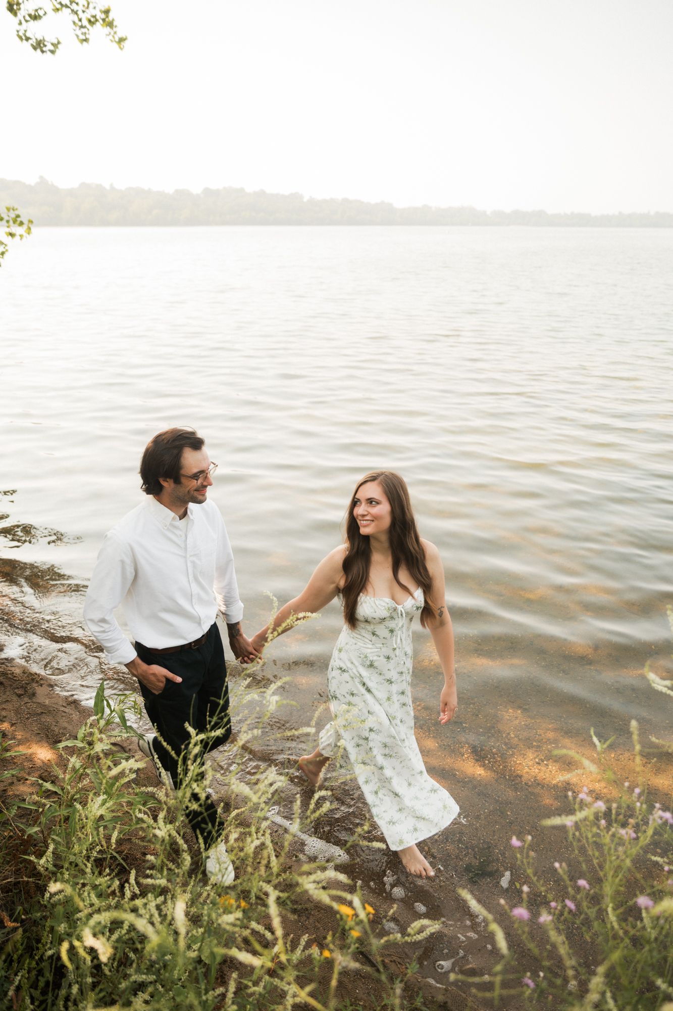A couple holds hands and walks along a lakeside shore, the woman in a floral dress smiling at her partner.