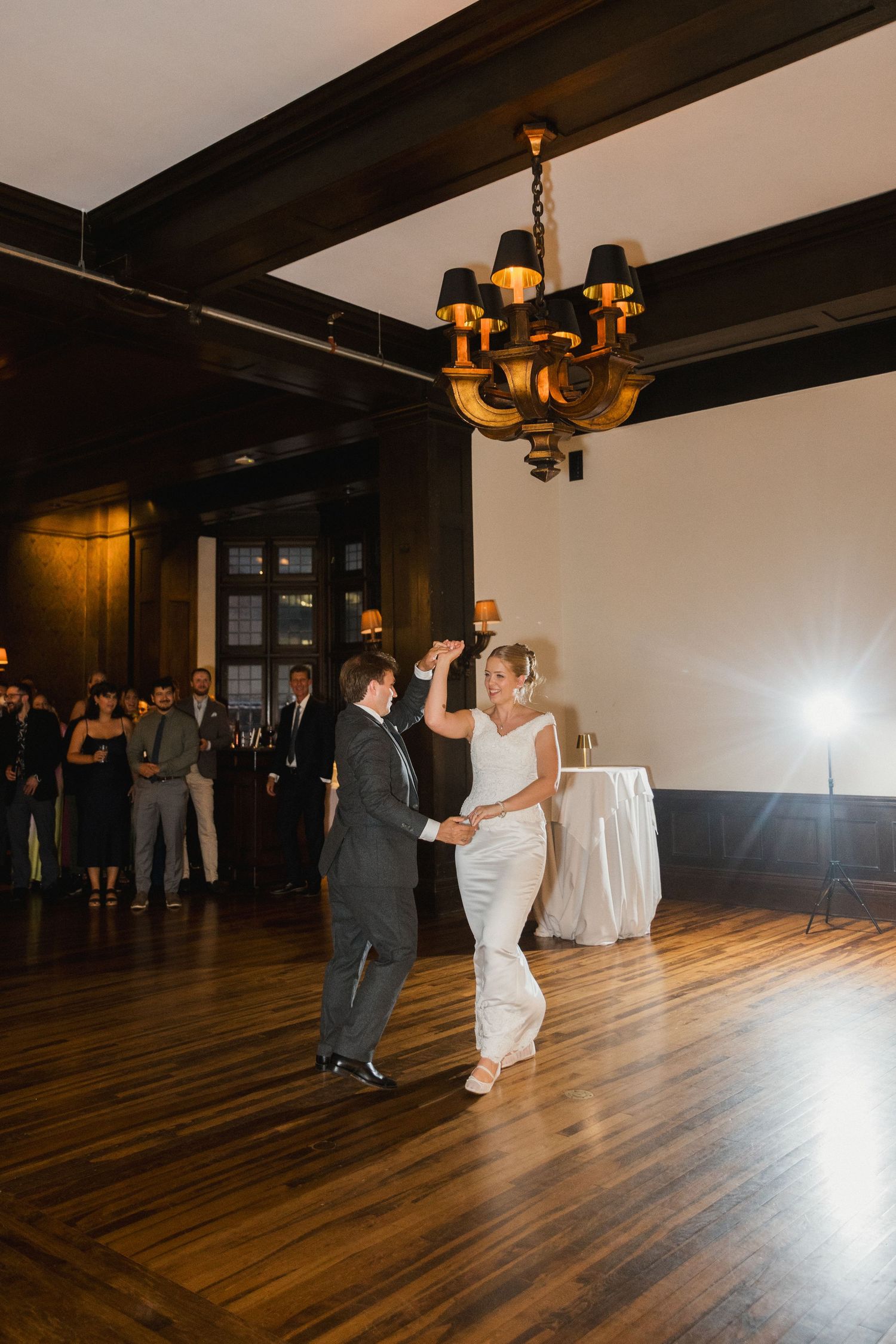 Bride and groom share first dance under chandelier in elegant dark wood ballroom with guests watching.
