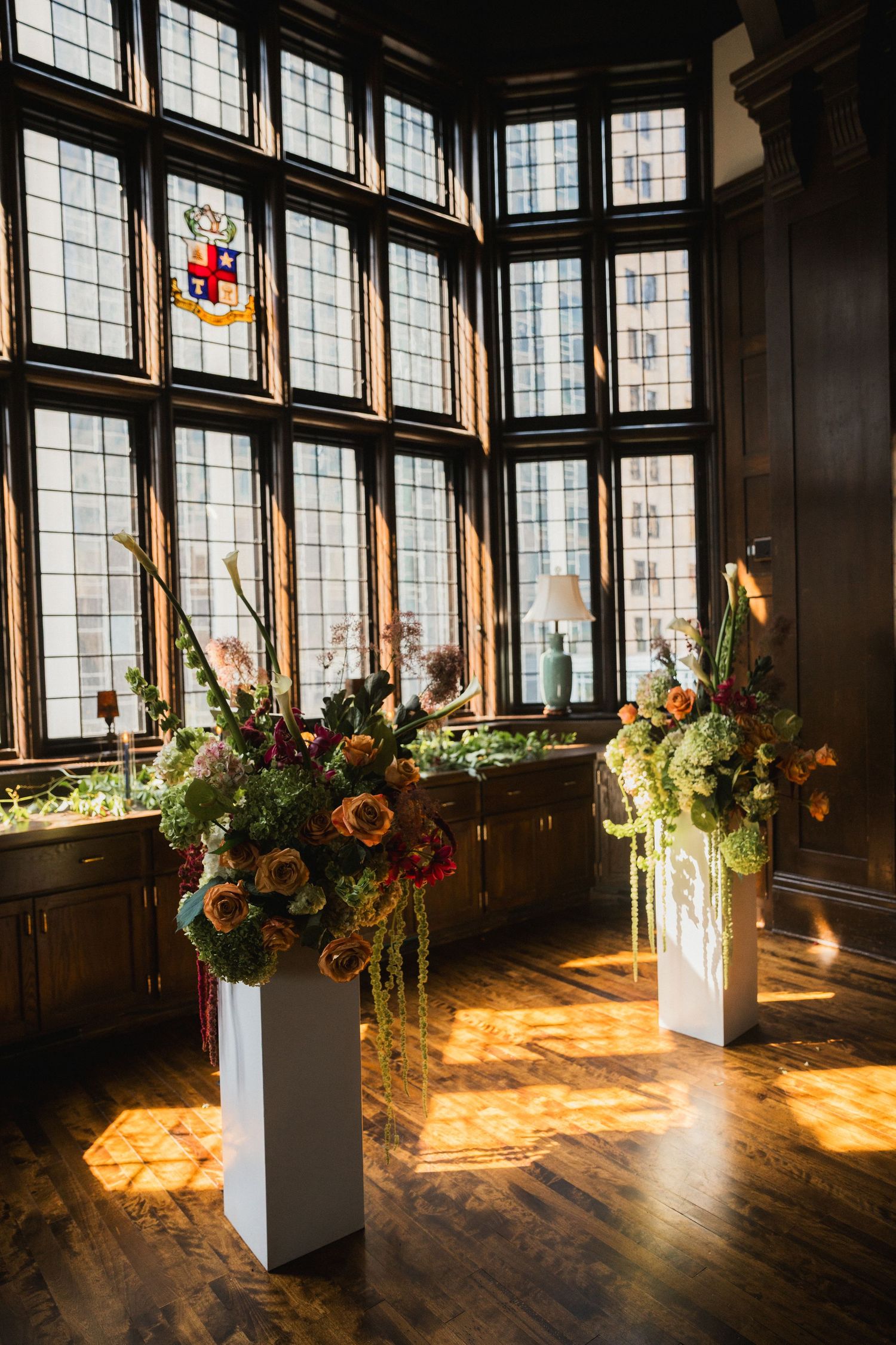Elegant wedding ceremony altar with lush floral arrangements on white pedestals in sunlit historic venue.