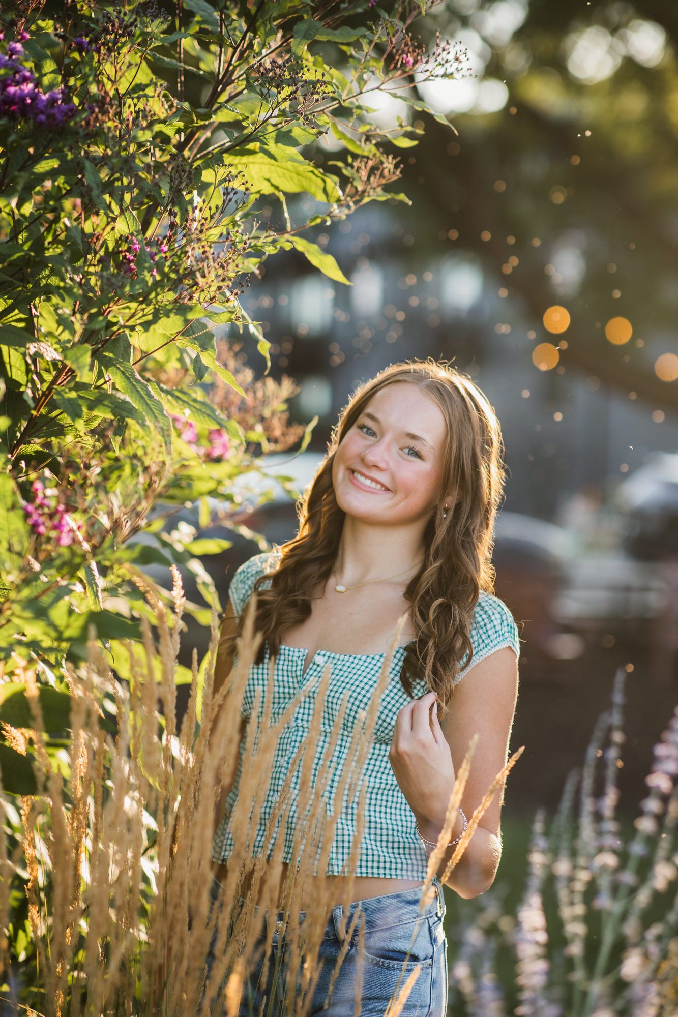 Young woman in teal gingham top smiles among lush garden plants in warm golden sunlight.