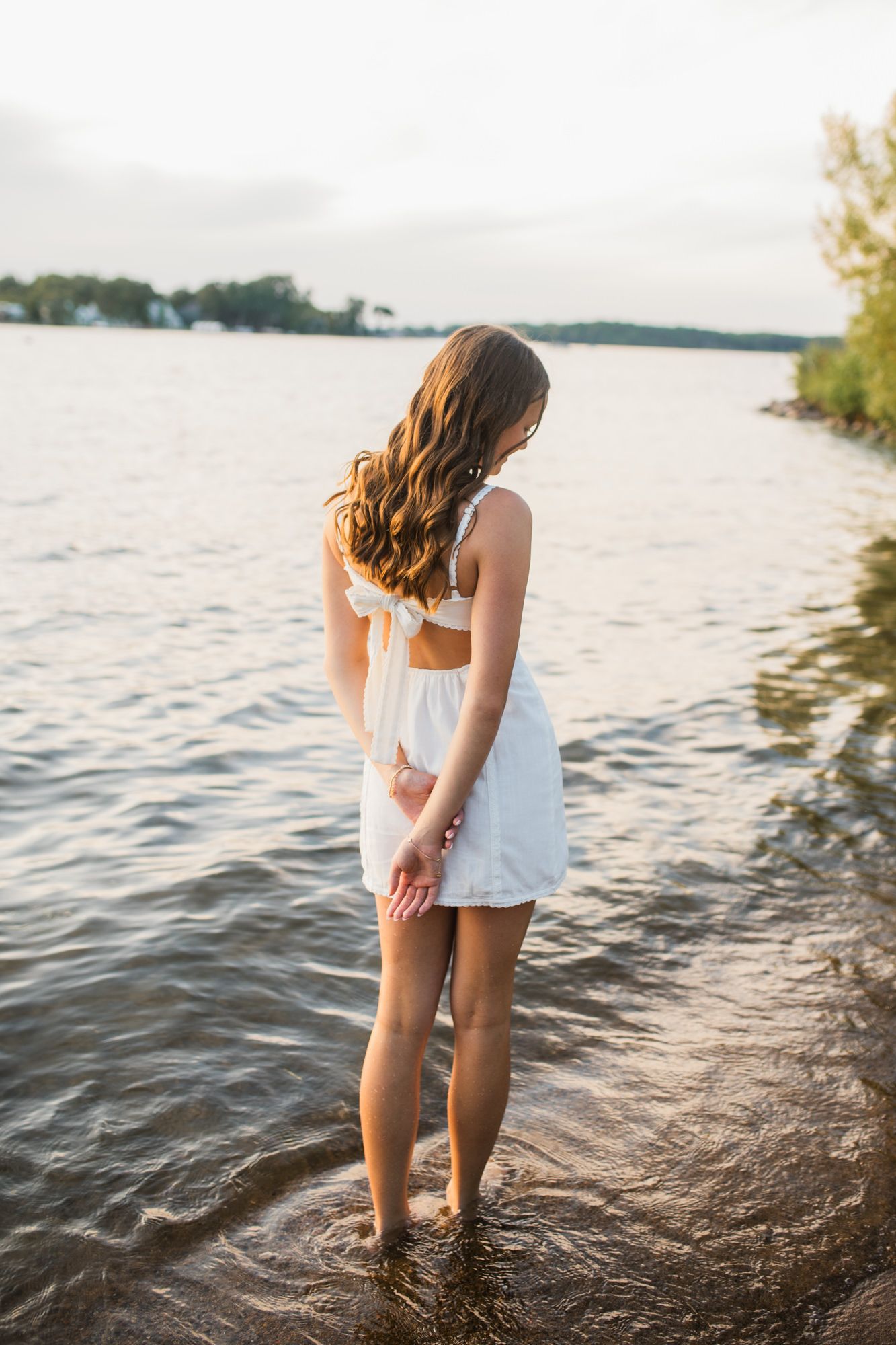 Young woman in white dress standing in shallow lake water at sunset, viewed from behind with long wavy hair.