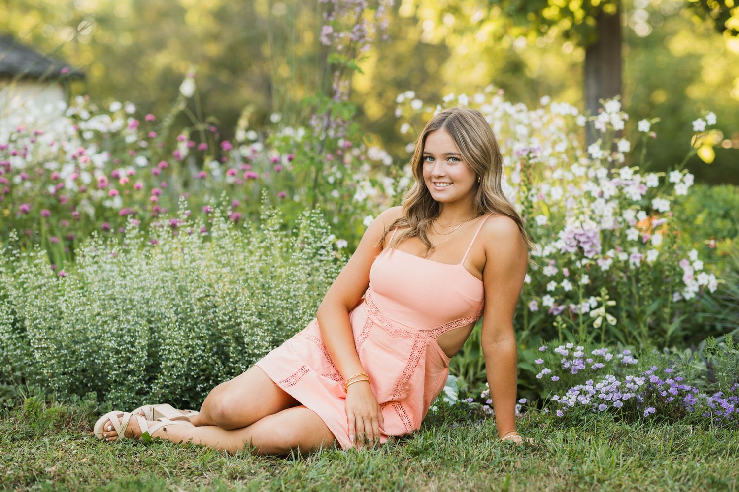 Young woman in pink dress sitting on grass surrounded by colorful wildflowers in a sunny garden.