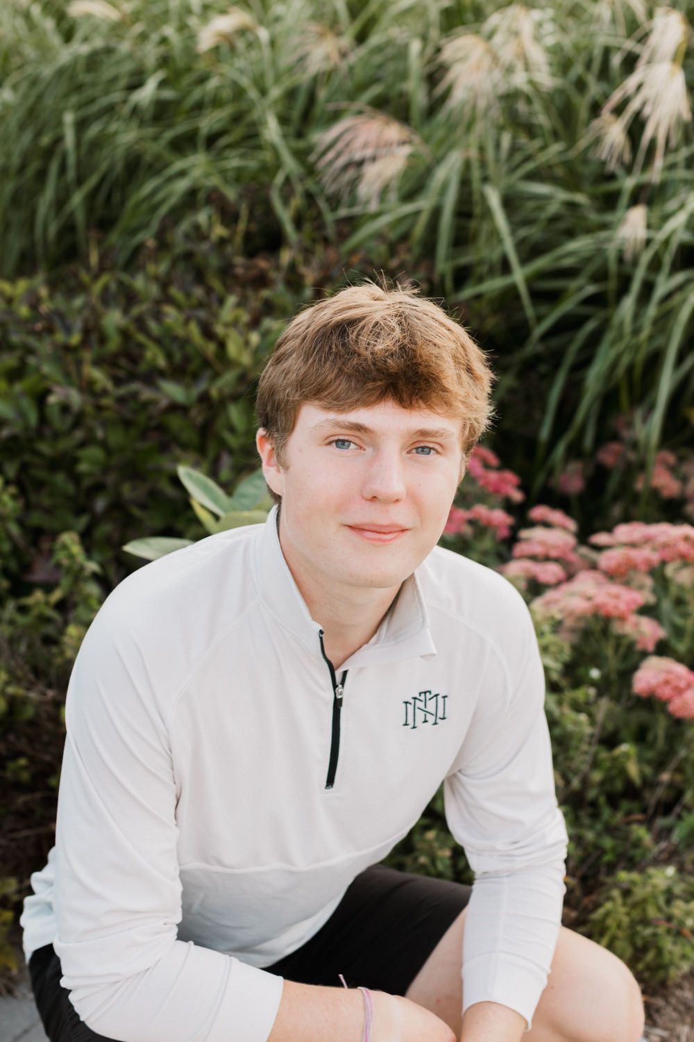 Young person in a white quarter-zip pullover poses outdoors against floral garden backdrop.