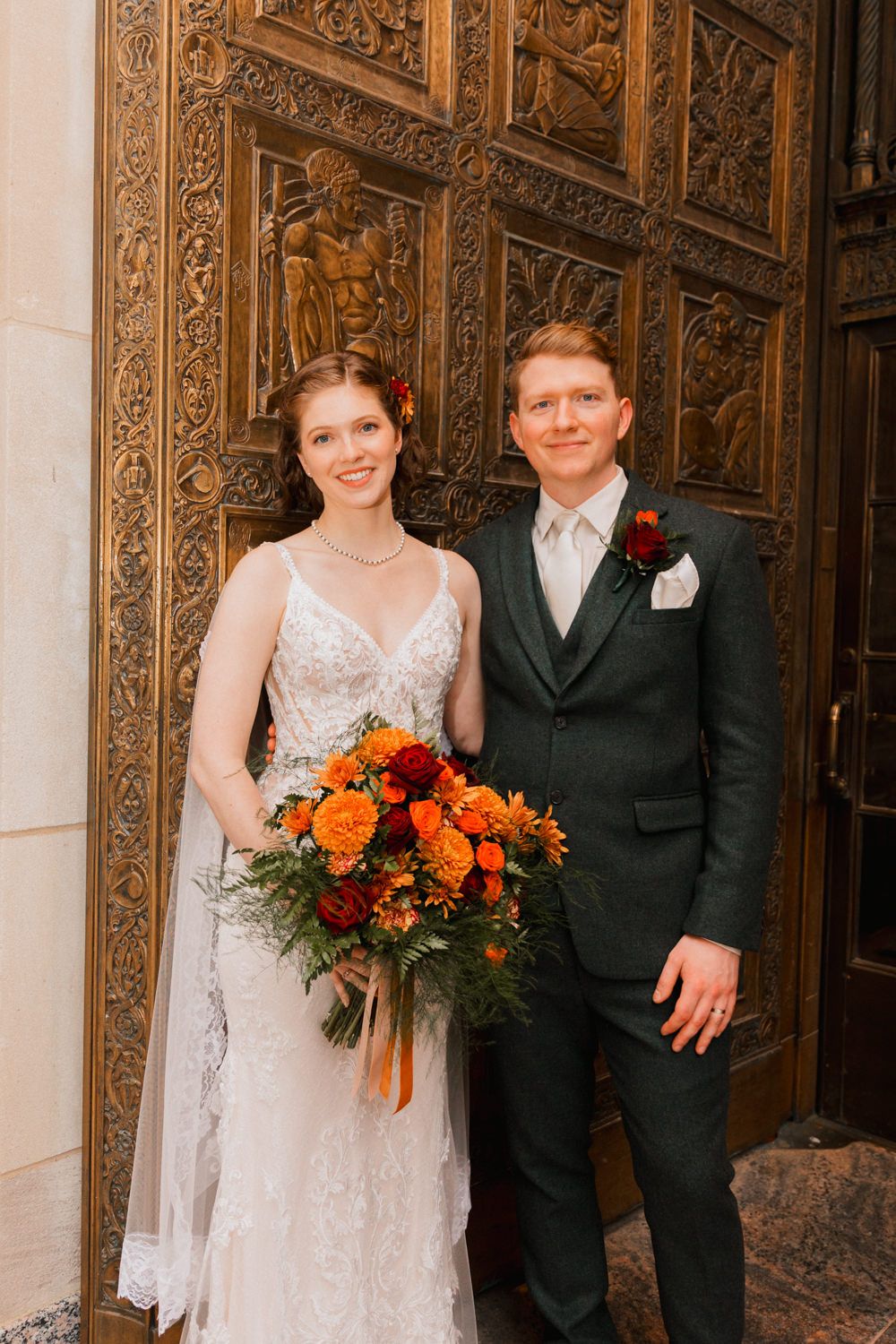 A couple poses in front of ornate wooden doors with an orange and red floral bouquet.