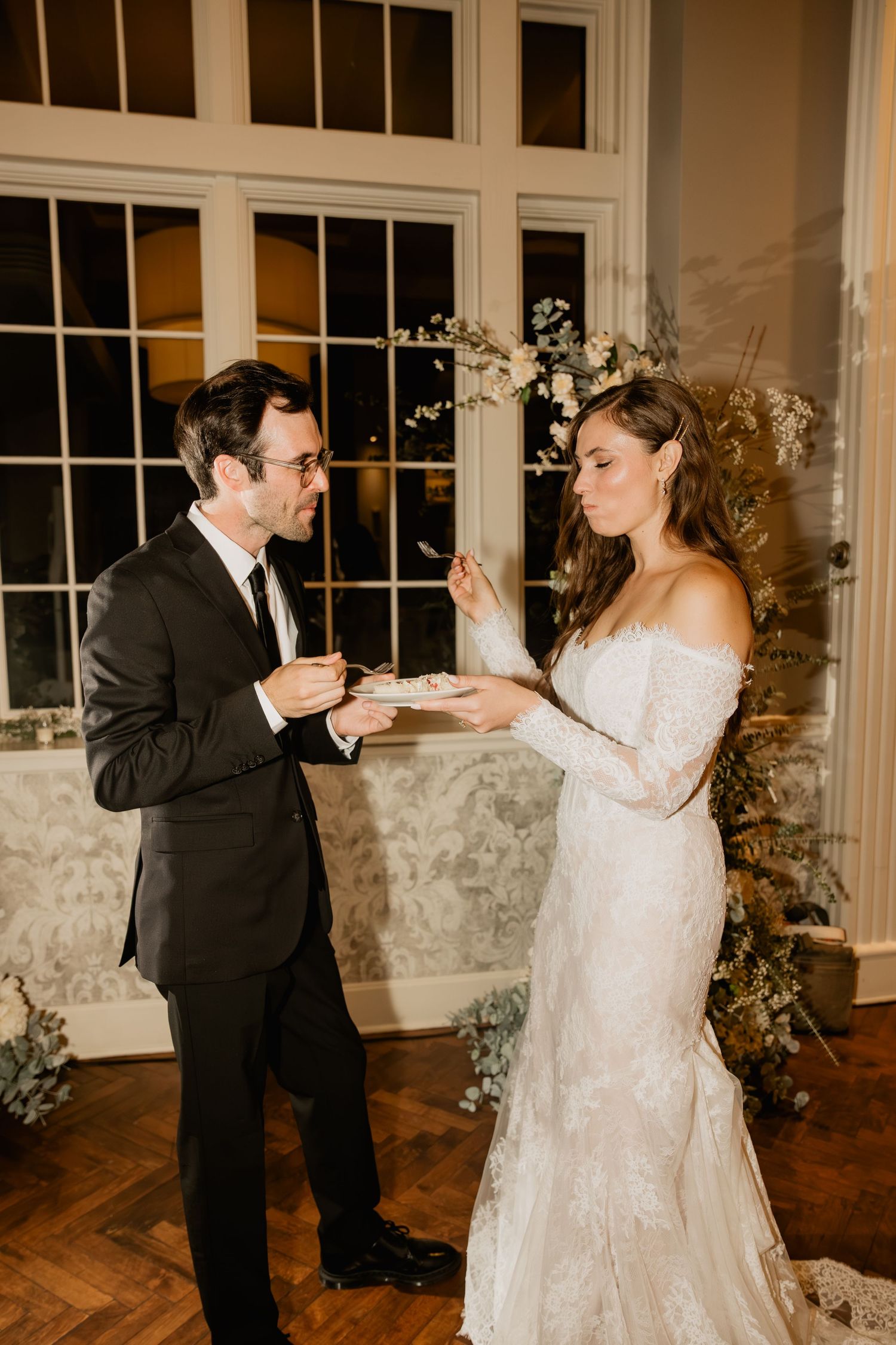 Bride and groom sharing wedding cake at elegant indoor reception, bride in lace off-shoulder gown, groom in black suit.
