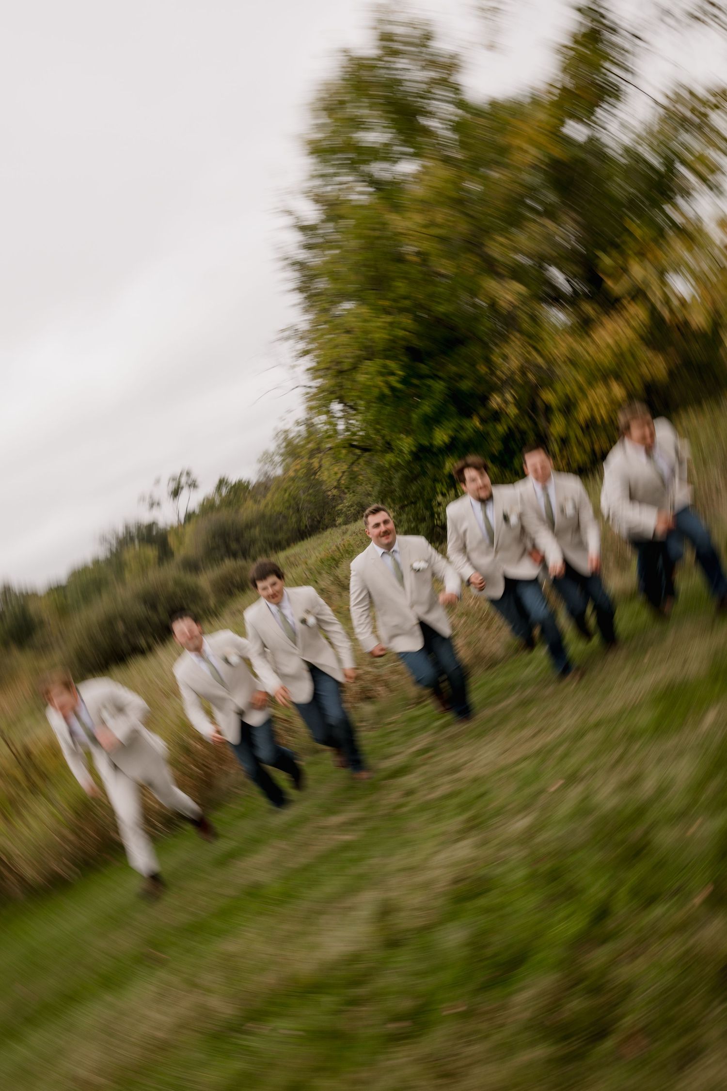 Groomsmen in tan blazers and jeans run through a green field in a motion-blur wedding photo.
