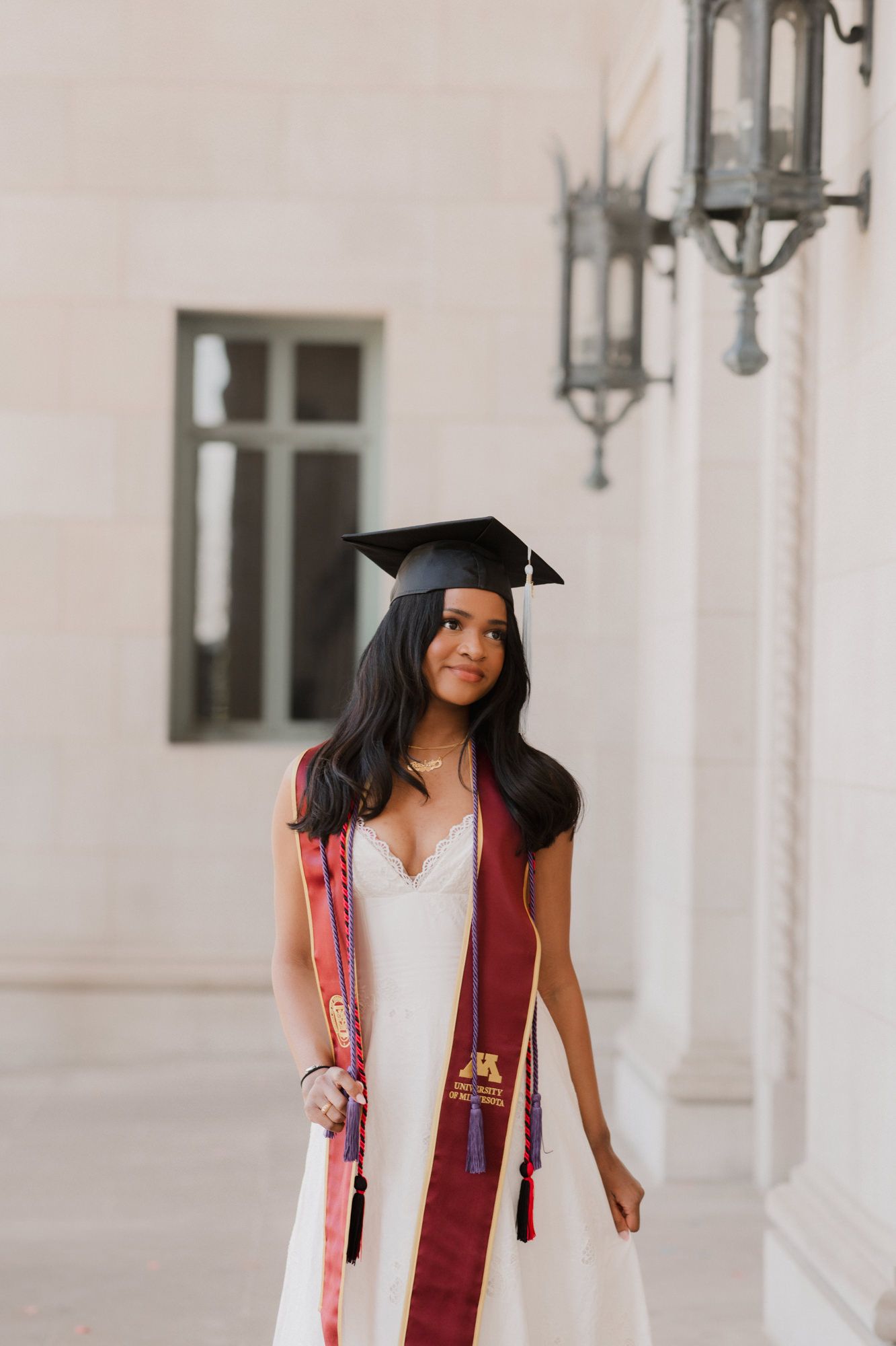 Young woman in graduation cap and maroon stole over white dress poses outdoors at a university building.